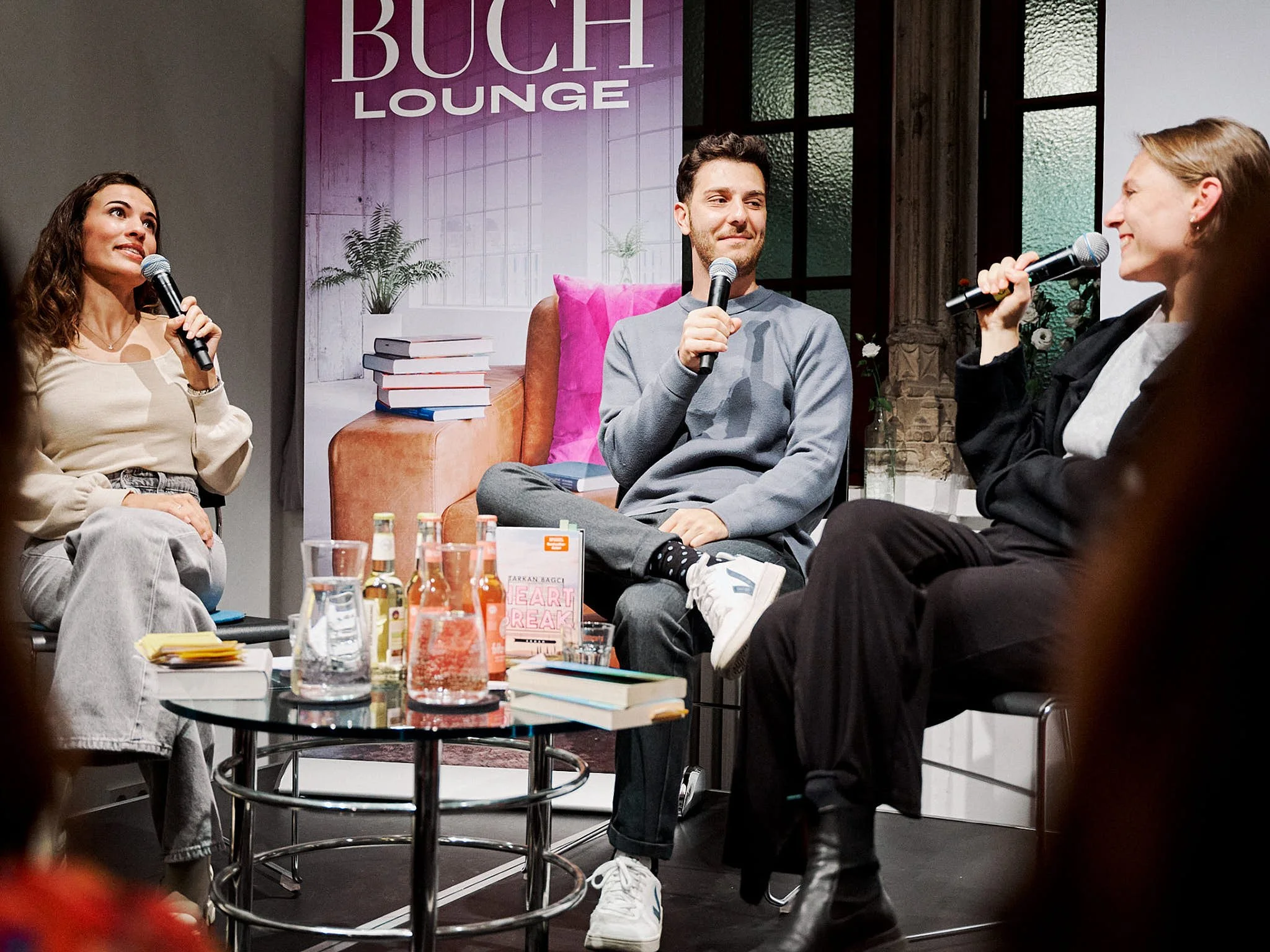 Drei Menschen bei einem Gespräch auf einer Bühne, mit Mikrofonen in der Hand, vor einem Banner mit der Aufschrift 'BUCH LOUNGE', in einem stilvollen Raum mit großen Fenstern und Dekoration.