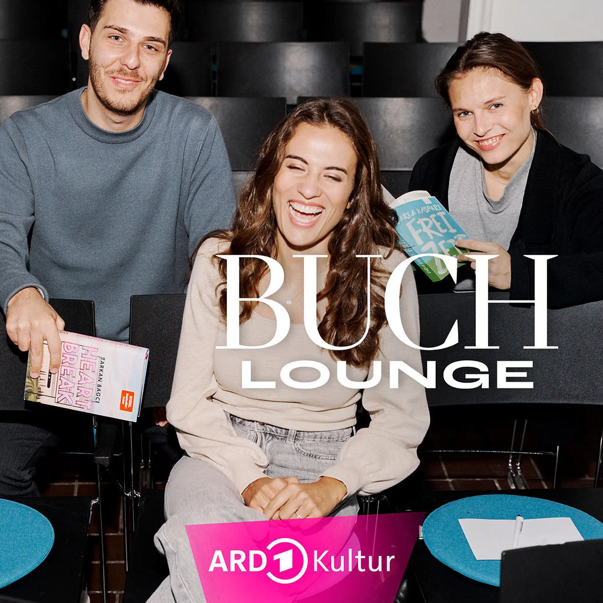 Drei lächelnde Menschen sitzen in einem dunklen Raum, vermutlich in einem Theater oder Kino. Zwei Frauen und ein Mann, die Frau in der Mitte lacht herzlich. Es liegt eine Zeitschrift oder ein Buch in der Hand des Mannes, und die Frau rechts hält ein weiteres Buch. Der Text "BUCH LOUNGE" und "ARD Kultur" ist auf dem Bild.