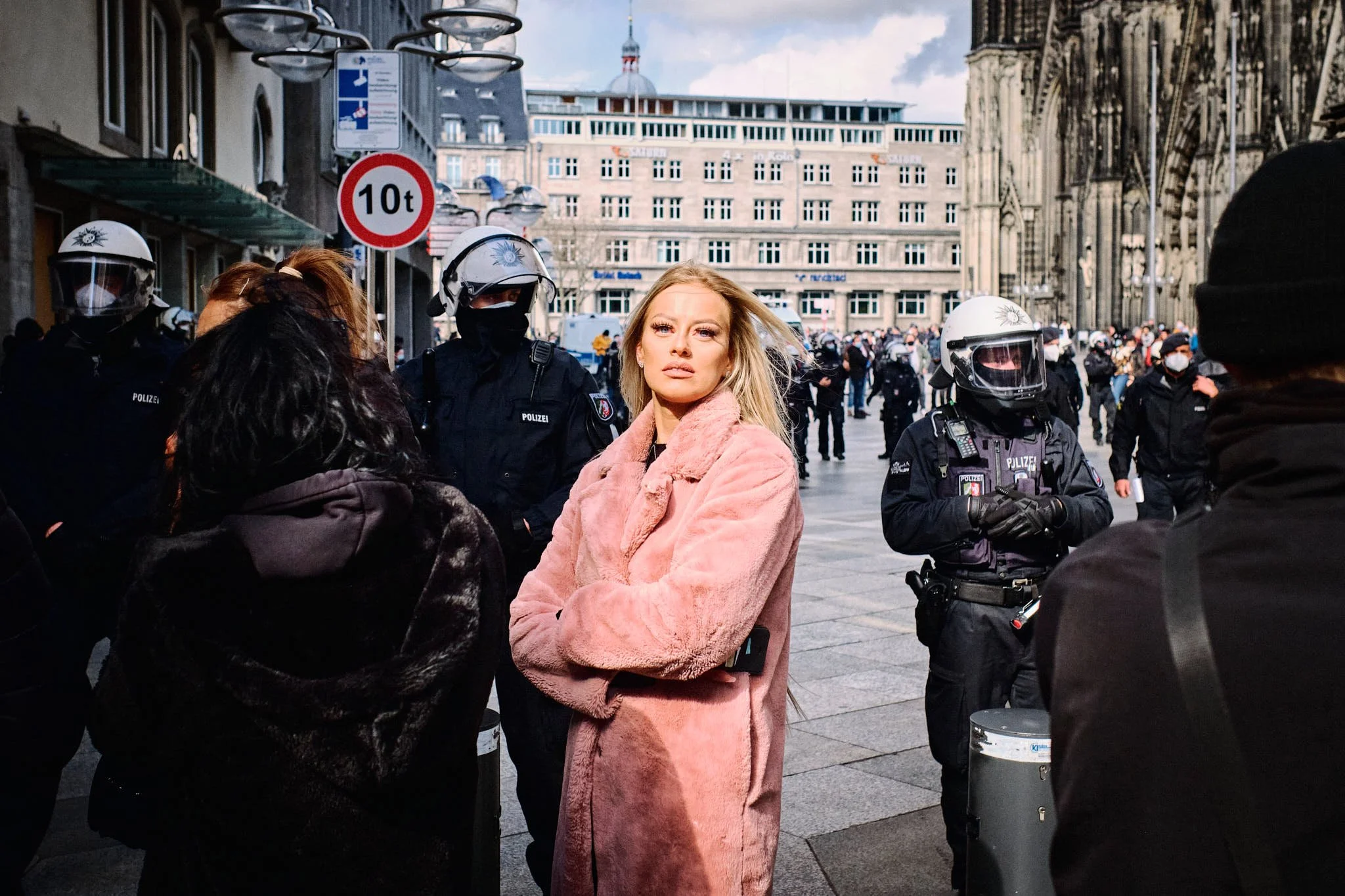 Eine Frau in einem pinken Mantel steht inmitten einer Menschenmenge, umgeben von Polizei mit Helmen, in einer Stadt vor historischen Gebäuden.