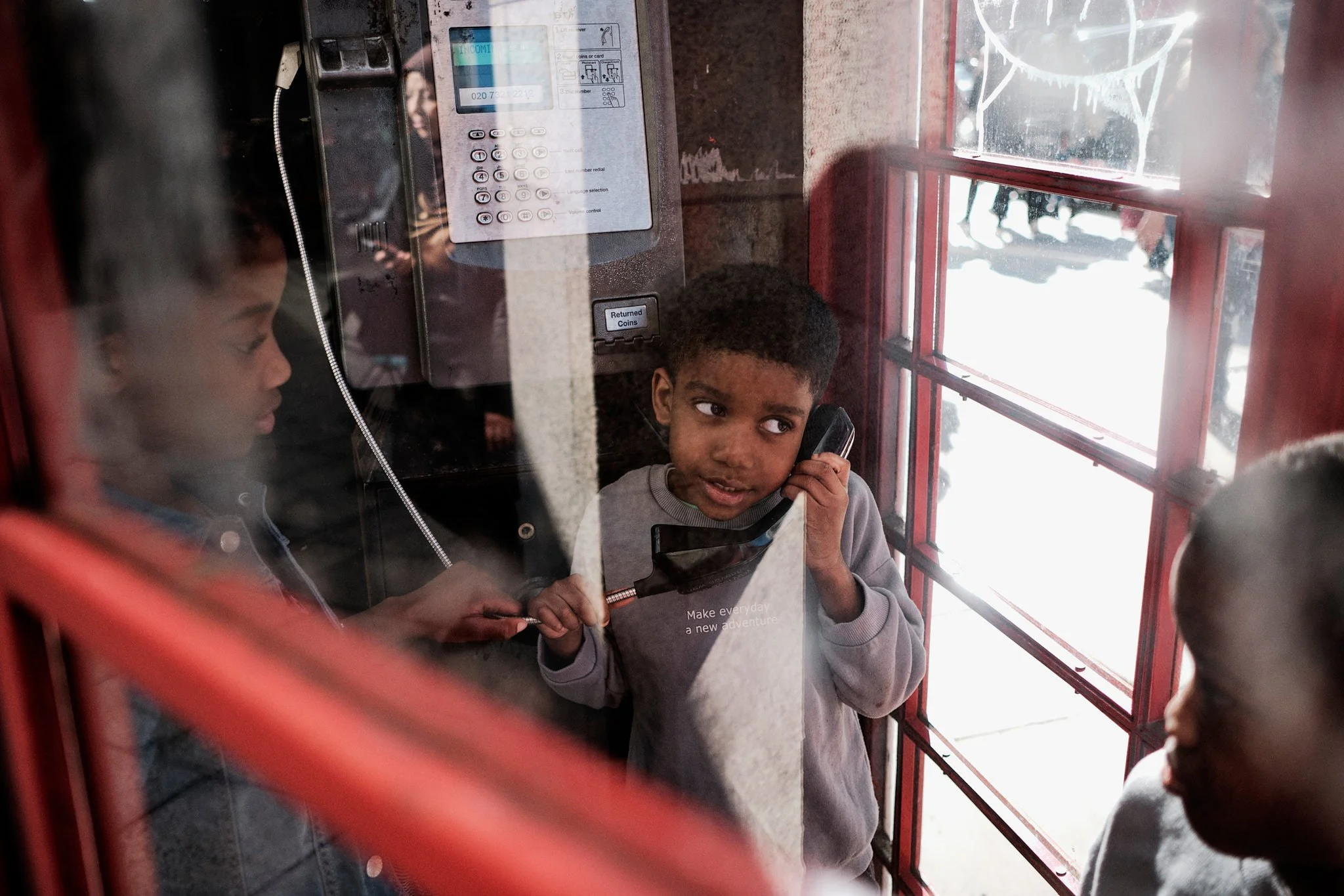 Kinder im Telefongespräch in einer Telefonzelle, eine der Kinder hält das Telefon, während die anderen beobachten.