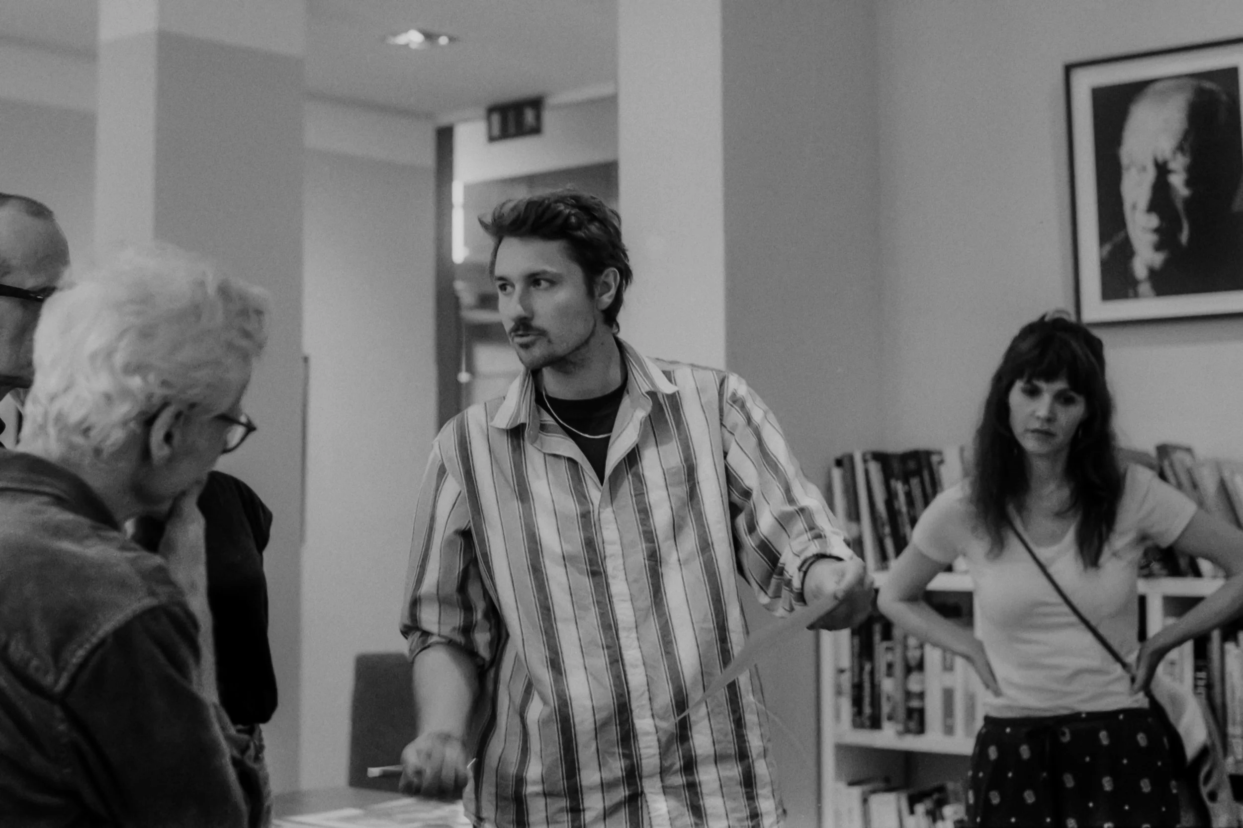 Drei Menschen unterhalten sich in einem Raum mit Büchern und einem Foto eines Mannes an der Wand, Schwarzweißbild.