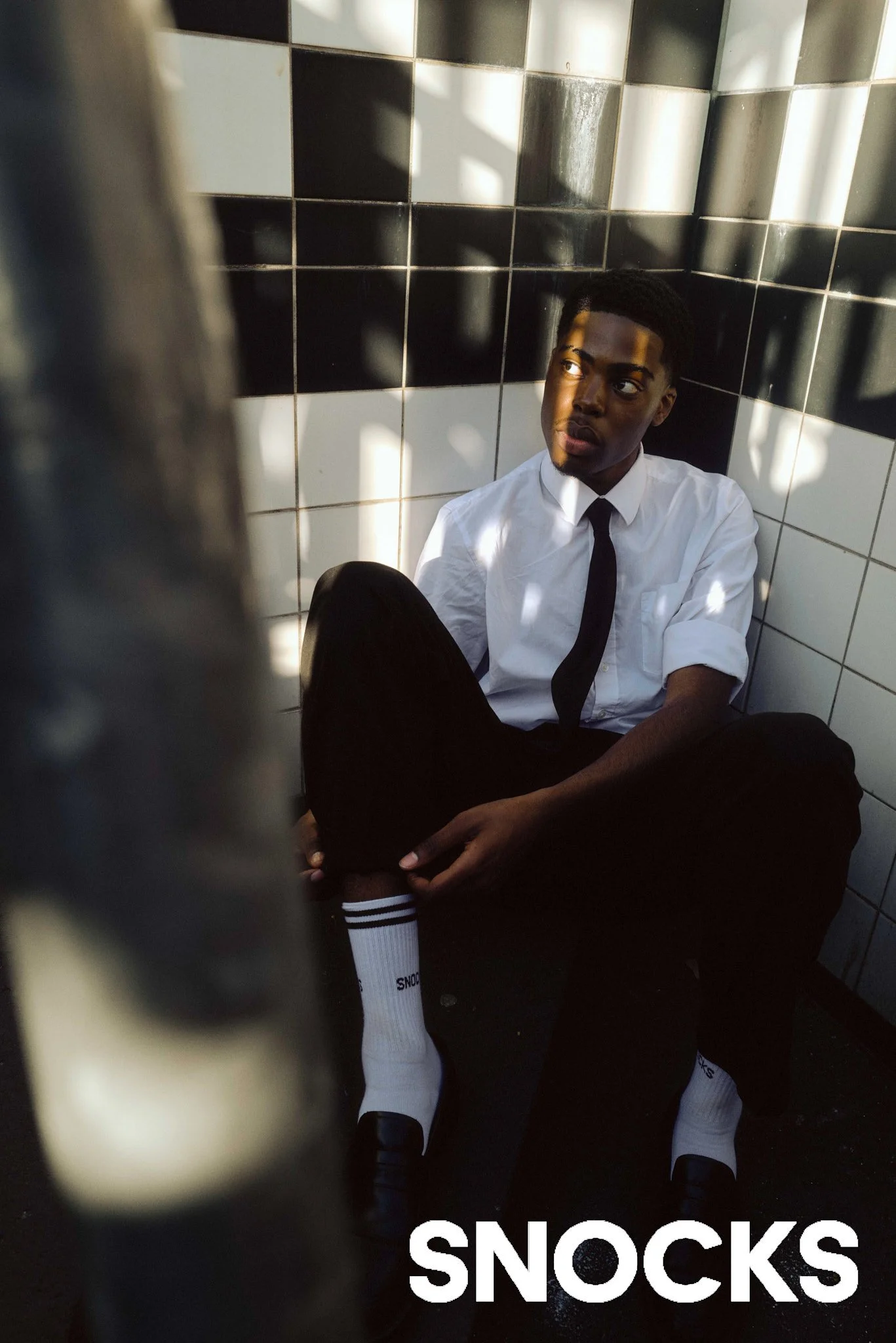 Ein junger Mann in einem weißen Hemd, schwarzer Krawatte und schwarzen Hosen sitzt auf dem Boden in einer Ecke mit schwarz-weißen Kacheln. Er trägt weiße Socken mit schwarzer Spitze, die mit "SNOCKS" beschriftet sind. Der Mann schaut nach oben, während Schatten den Raum durchdringen.