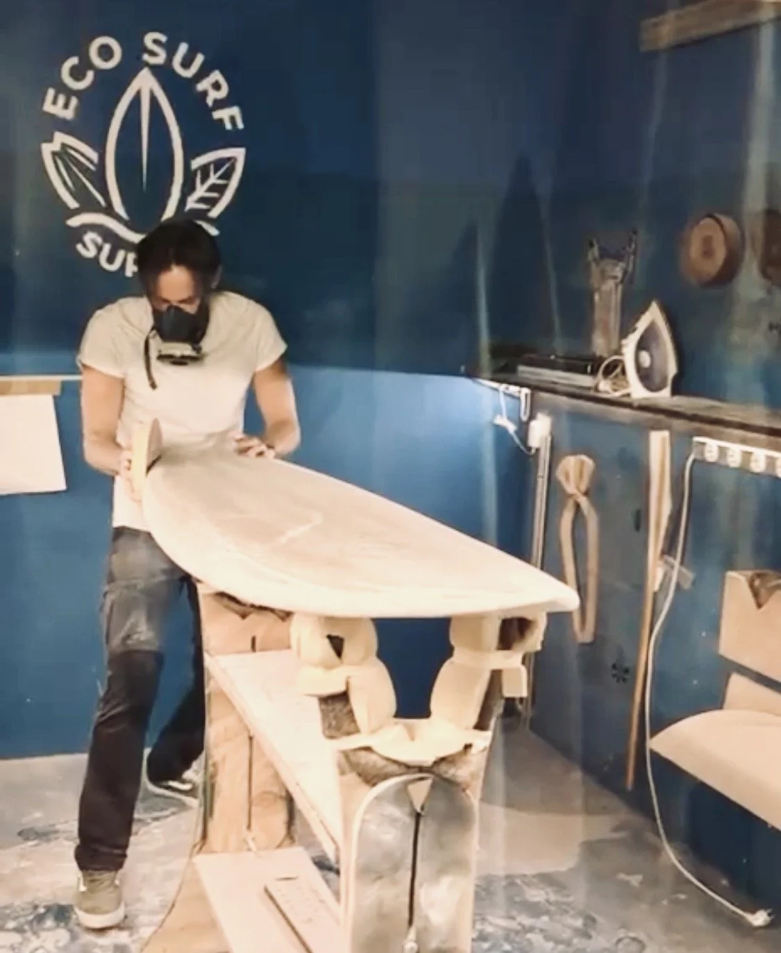 A person with a mask sanding a wooden surfboard in a workshop with a 'ECO SURF' logo on the blue wall.