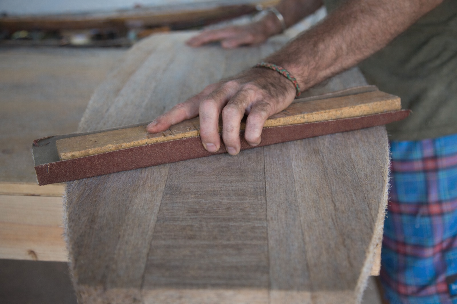 A person's hand sanding a wooden surface with a sanding block in a woodworking shop.