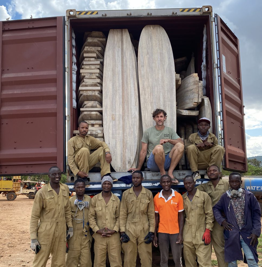 A group of men, some in work clothes and some in casual clothes, pose in front of a large shipping container filled with wooden surfboards and other wooden objects. Some men are sitting on the edge of the container, while others stand in front.