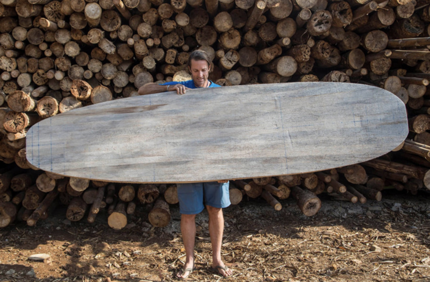 A man holding a large wooden surfboard in front of a stack of logs.