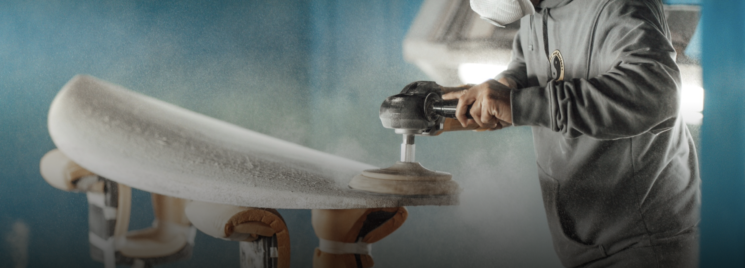 A person wearing gloves and a gray jacket is polishing a surfboard with an electric tool, creating a spray of dust or polish.