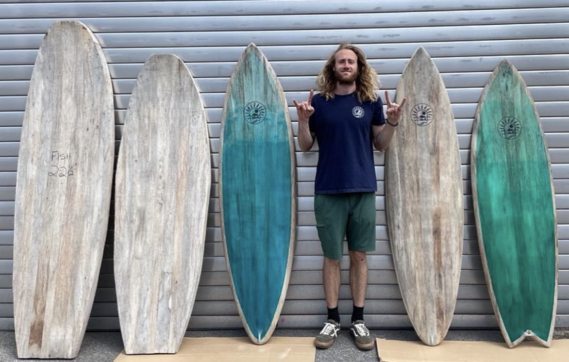 A man with long curly hair and a beard, wearing a navy blue t-shirt and green shorts, standing between five vintage wooden surfboards against a metal wall, making a rock and roll hand gesture with both hands.