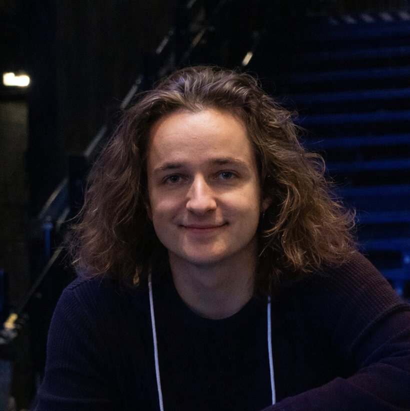 Ohjaaja Kirmo Komulainen istuu pimeässä tilassa, taustalla ritiläportaita ja sinistä valaistusta.