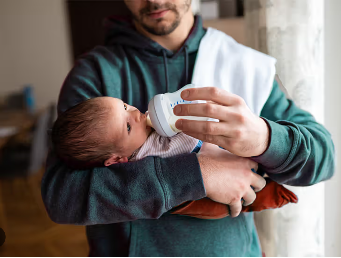 A man holding a baby while feeding them with a baby bottle indoors.