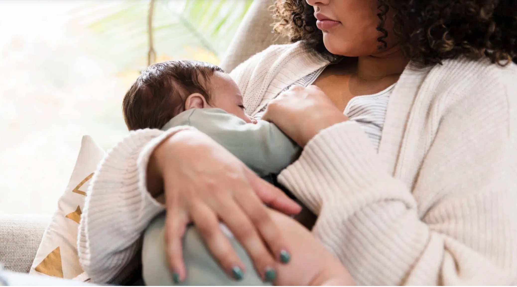 A woman with curly hair holds a baby close to her chest, nursing the baby while sitting on a sofa in a bright room with a window in the background.