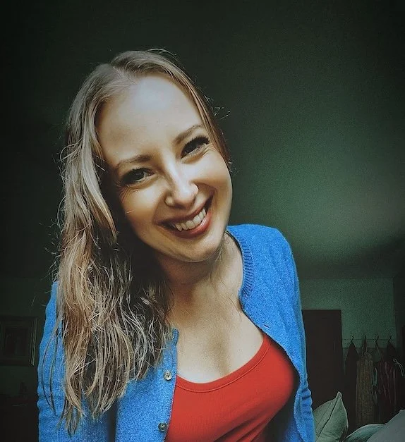 A young woman with long, wavy light brown hair, smiling, wearing a red top and a blue cardigan, in a room with a green wall and some furniture in the background.