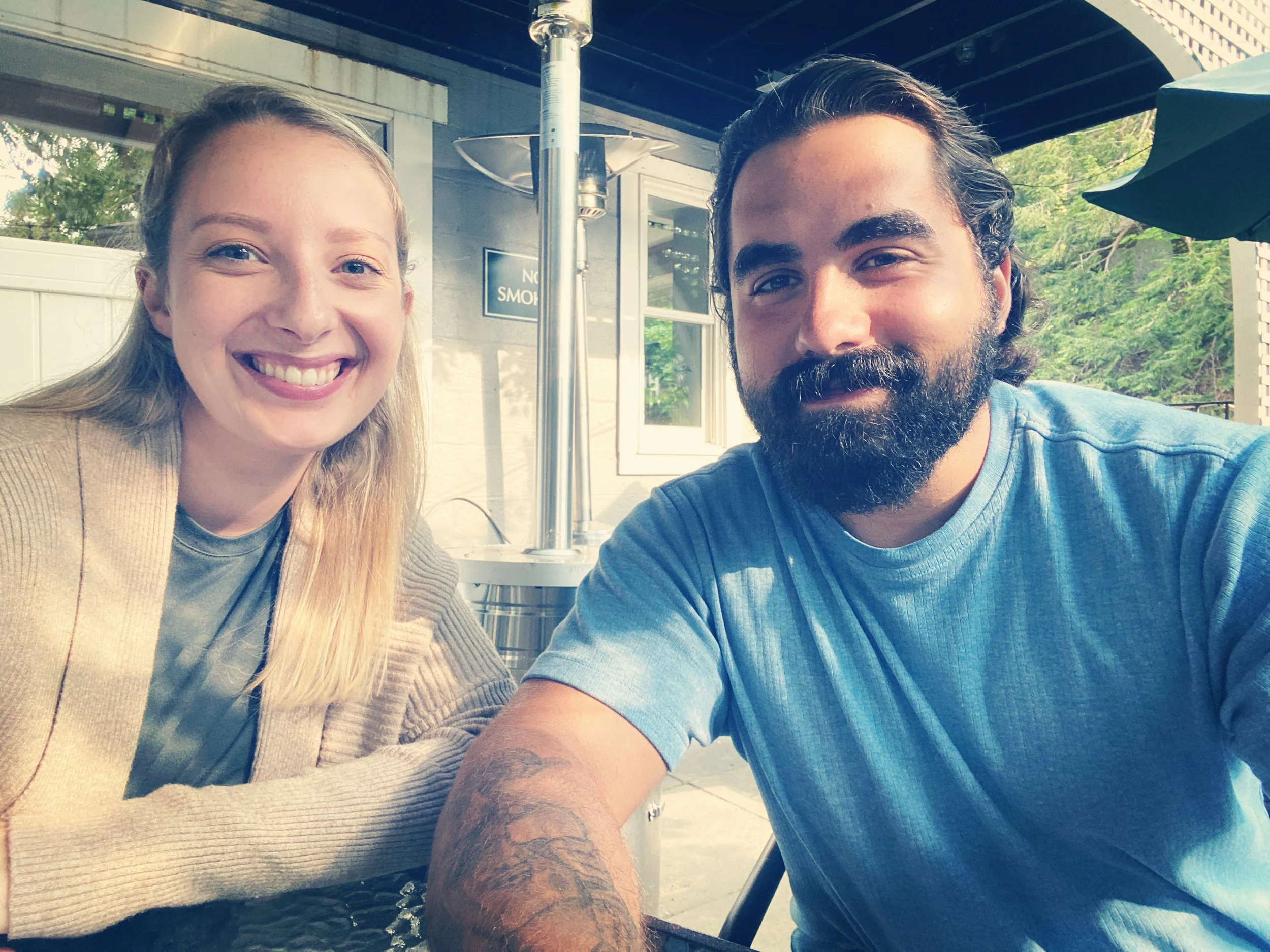 A smiling blonde woman and a bearded man with dark hair sitting at an outdoor table. They are taking a selfie with a patio, umbrella, and greenery in the background.