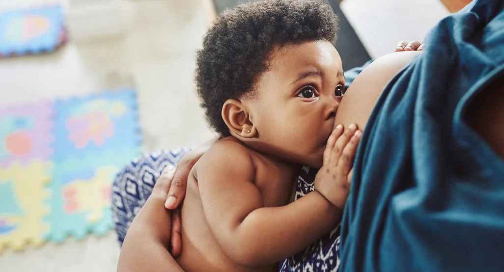 A baby breastfeeding from an adult, with a colorful play mat in the background.