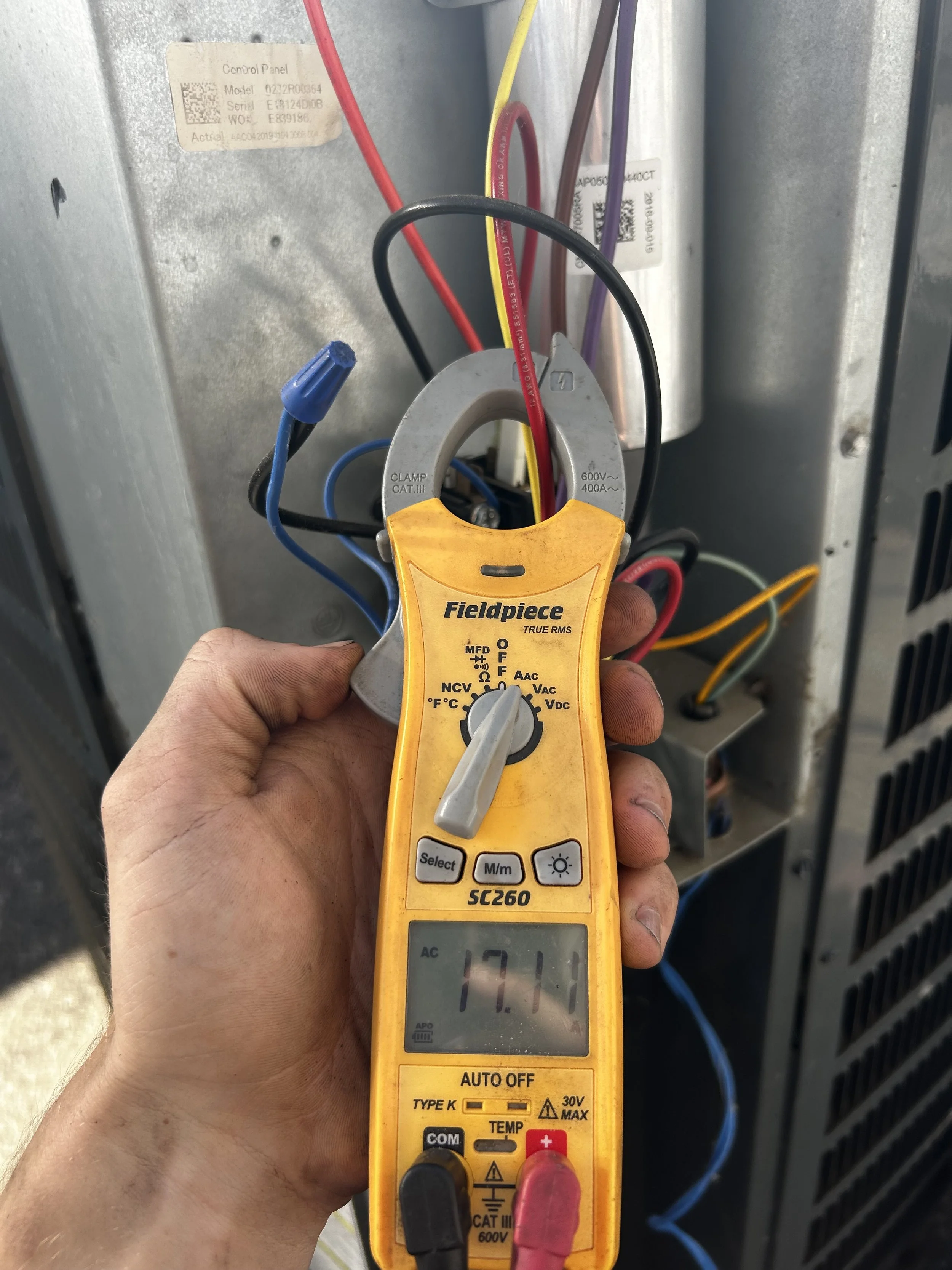 A digital multimeter measuring 171 volts AC in front of an electrical control panel with several colored wires connected.