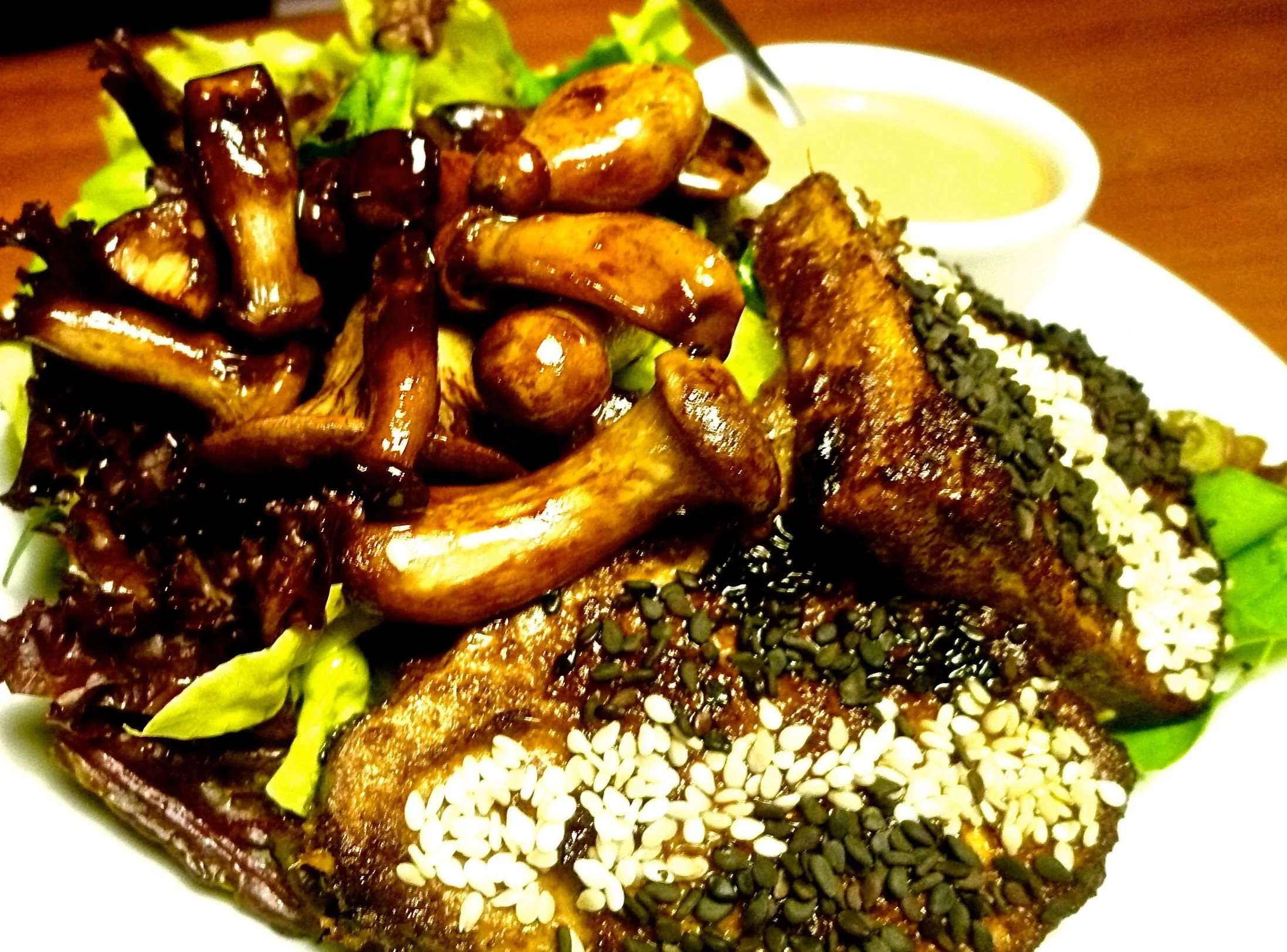 A plate of Asian-style food with oven baked tofu topped with black and white sesame seeds, stir-fried mushrooms, lettuce, and a side of dipping sauce.