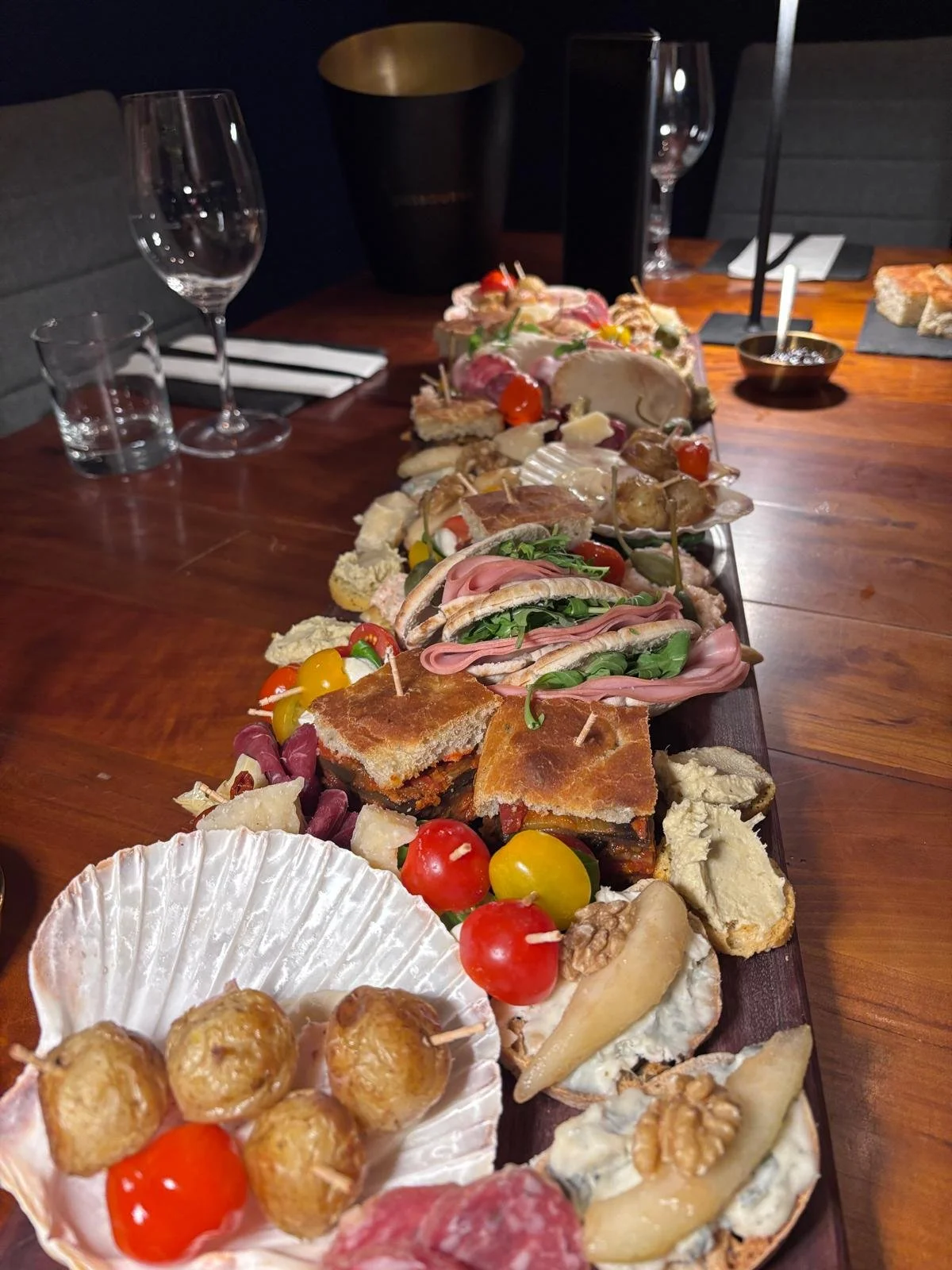 A long wooden serving board with an assortment of cheese, meats, vegetables, and appetizers. The table is set with wine glasses and napkins.