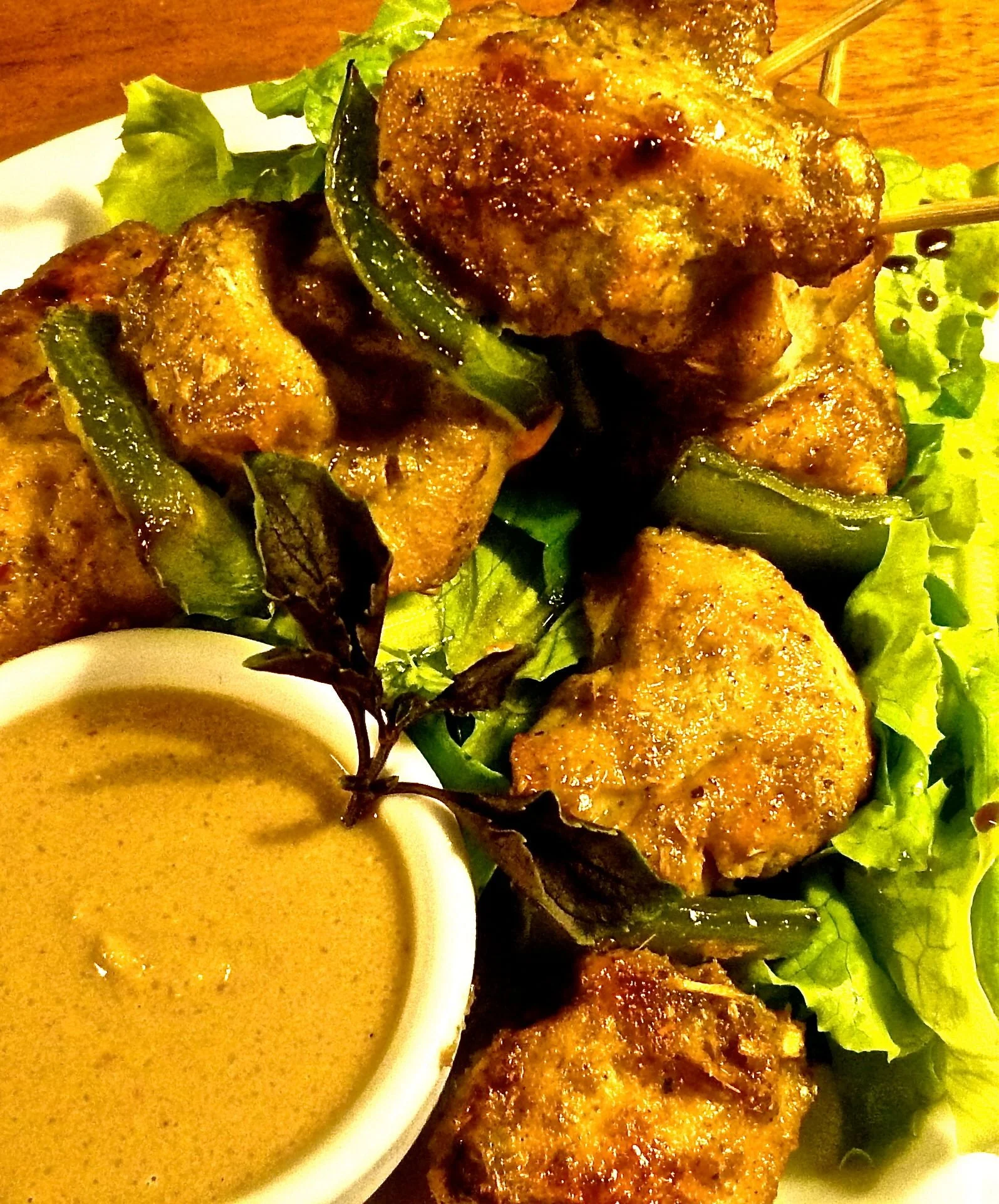 Chicken satay skewers garnished with green peppers, served with a side of dipping sauce and fresh leafy greens.