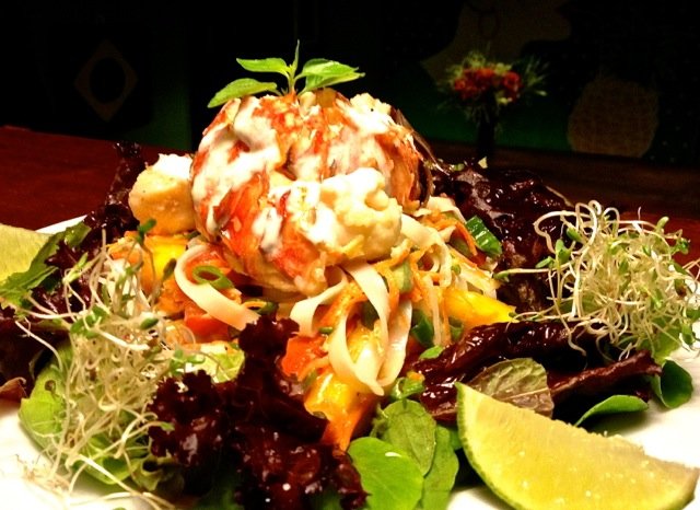 Lobster tail on a bed of mixed greens, shredded carrots, and sliced mango, garnished with microgreens and a lemon wedge.