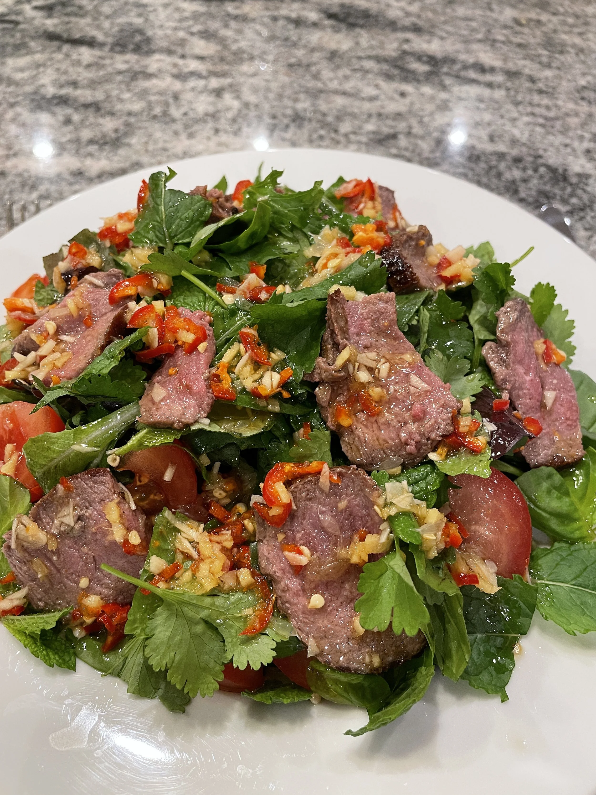 Steak salad with mixed greens, cherry tomatoes, red chili peppers, chopped garlic, and herbs on a white plate.