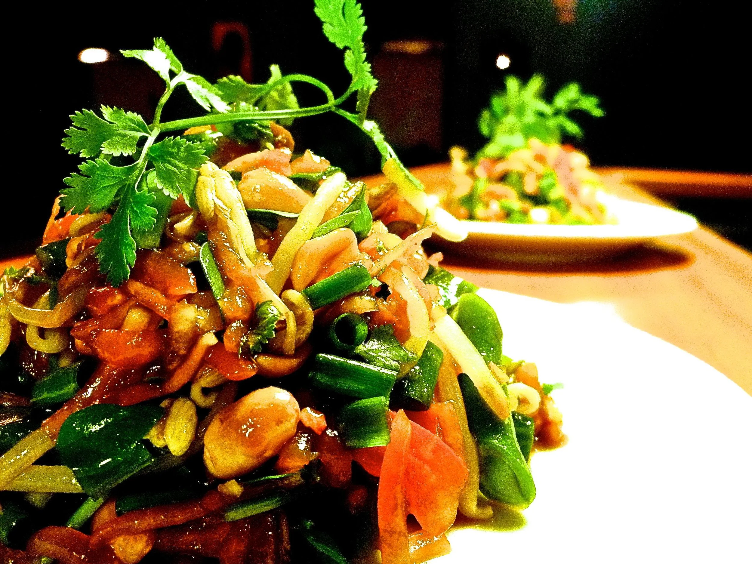 Close-up of a plate of Asian-style noodle salad garnished with coriander and green onions, with another similar dish in the background.
