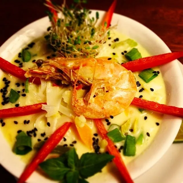 A bowl of Asian-style shrimp and noodle soup garnished with red bell pepper strips, chopped green onions, black sesame seeds, and a sprig of basil.