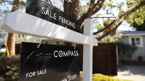 A real estate sign with 'Sale Pending' on top and 'Compass for Sale' at the bottom, located in a suburban neighborhood with trees and houses in the background.