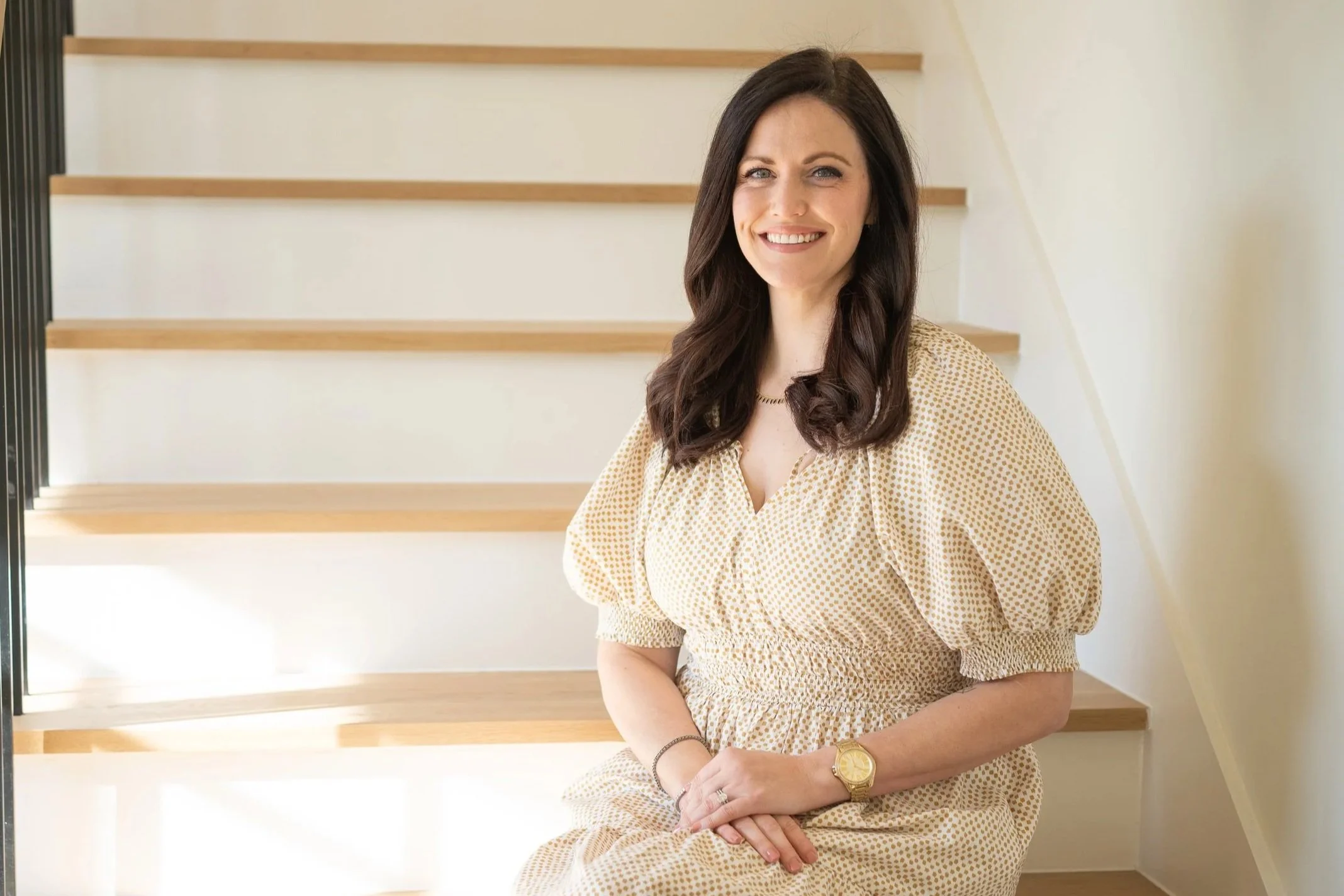 A woman with long dark hair, wearing a beige dress with small brown polka dots, sitting on wooden stairs, smiling at the camera. She is wearing a gold watch and a ring on her left hand.