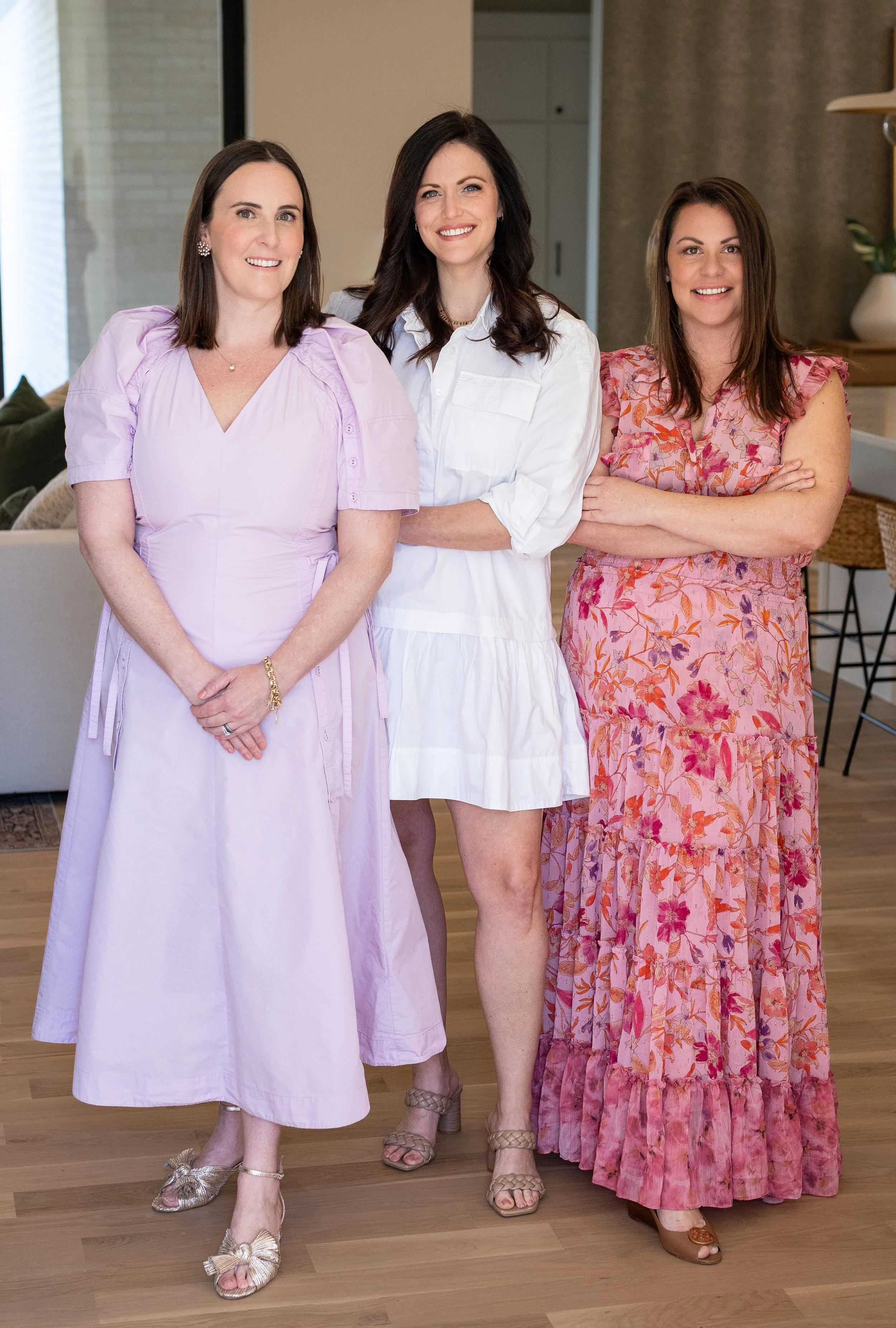 Three women standing inside a home, smiling at the camera. The woman on the left wears a light purple dress with puffed sleeves, the woman in the middle wears a white dress with rolled-up sleeves, and the woman on the right wears a pink floral dress with ruffled sleeves and a tiered skirt.
