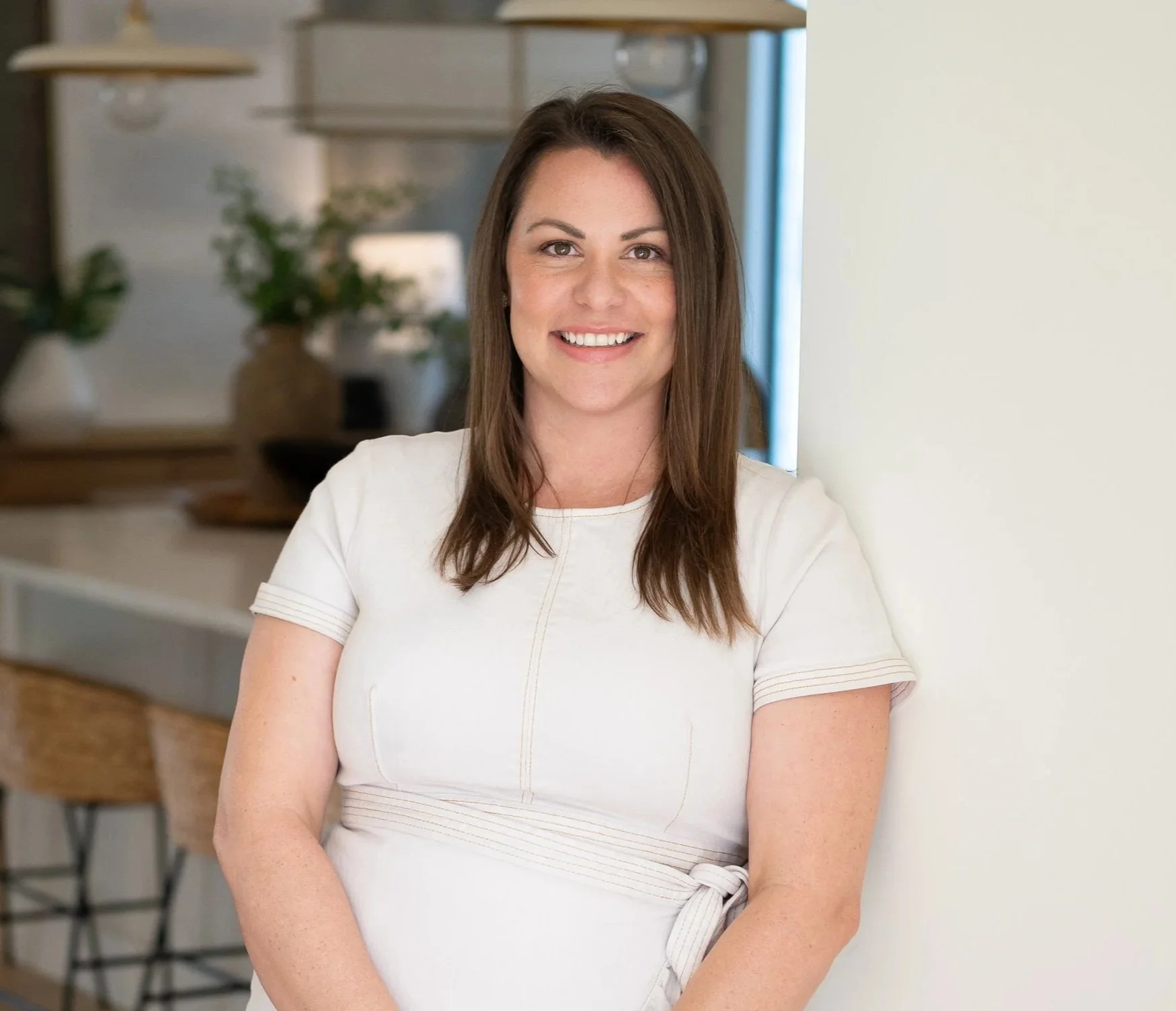 A woman with shoulder-length brown hair, smiling, wearing a white dress, standing indoors near a white wall with a kitchen in the background.