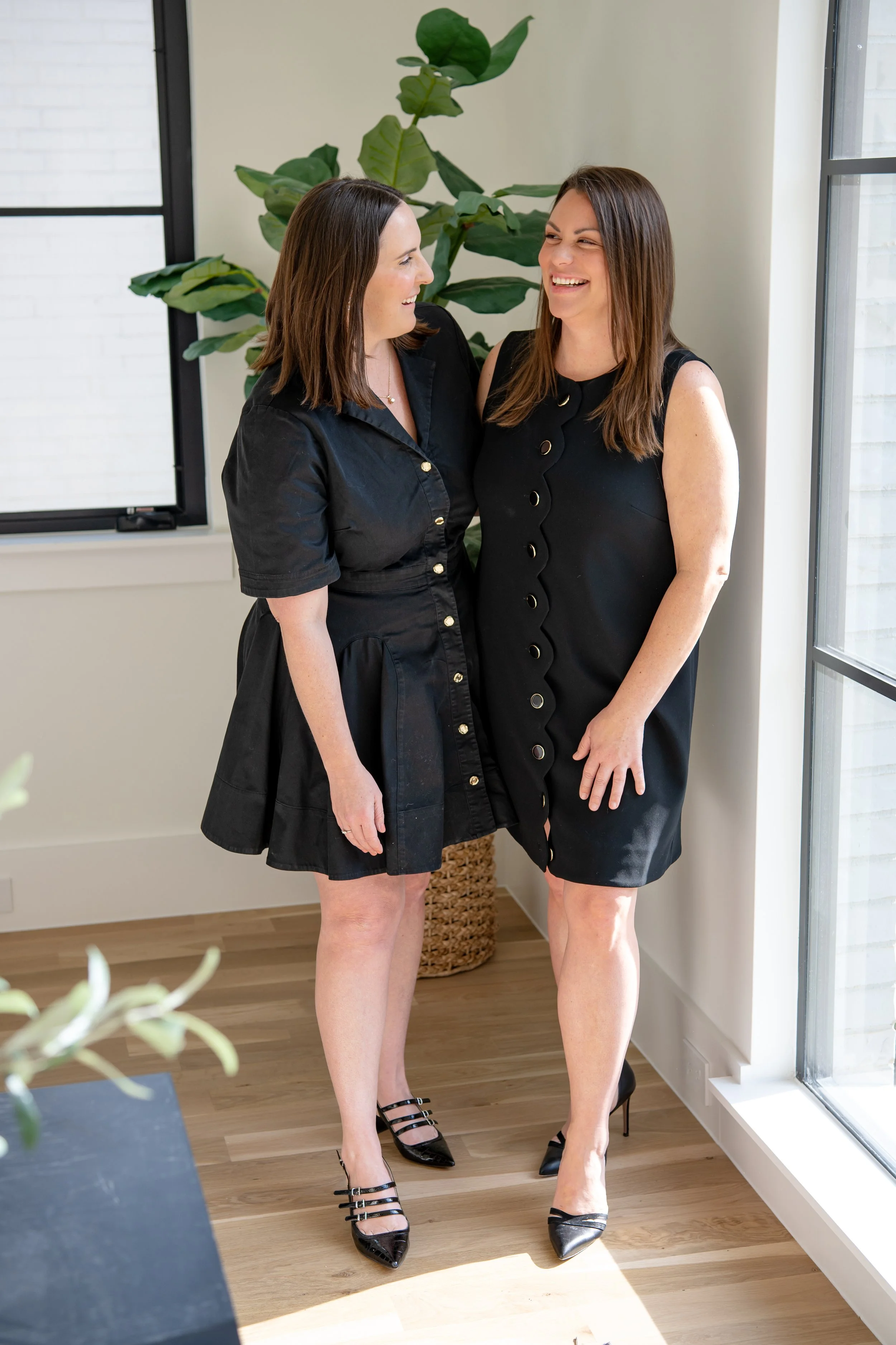 Two women in black dresses smiling and facing each other near a window and a large green plant in a bright room.