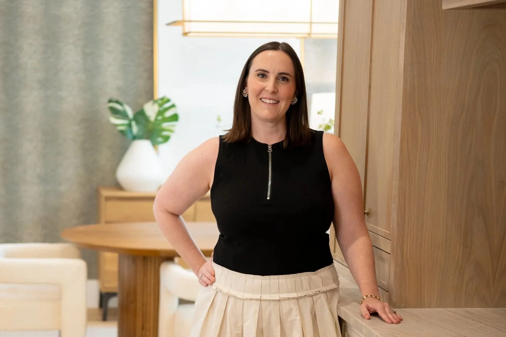 A woman smiling with brown hair, wearing a black sleeveless top with a zipper and a beige pleated skirt, standing indoors near wooden furniture and a white vase with green leaves.