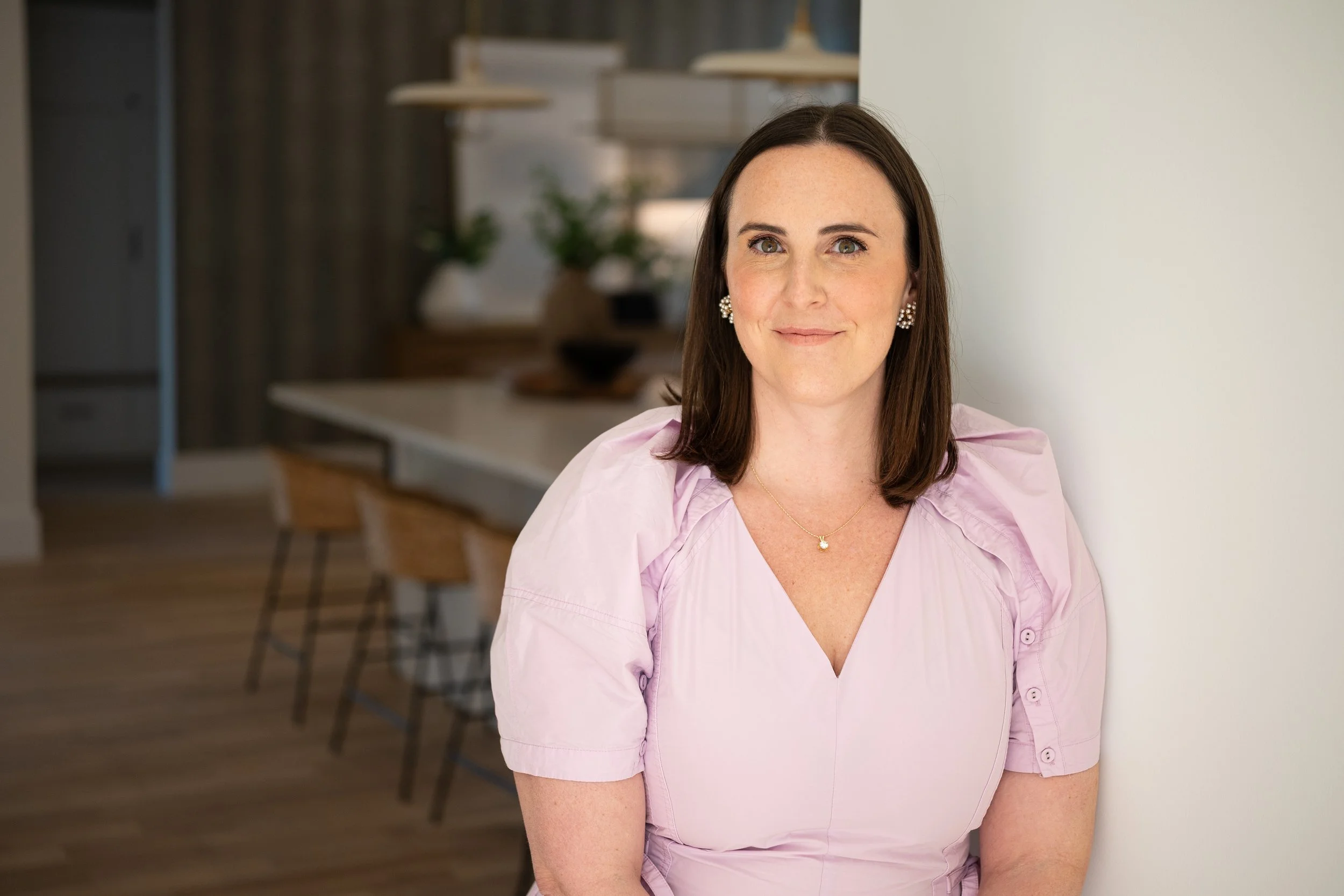 A woman with shoulder-length dark brown hair, wearing a light pink blouse, sitting against a white wall inside a modern home or apartment. She is smiling and wearing pearl earrings and a gold necklace with a small pendant.
