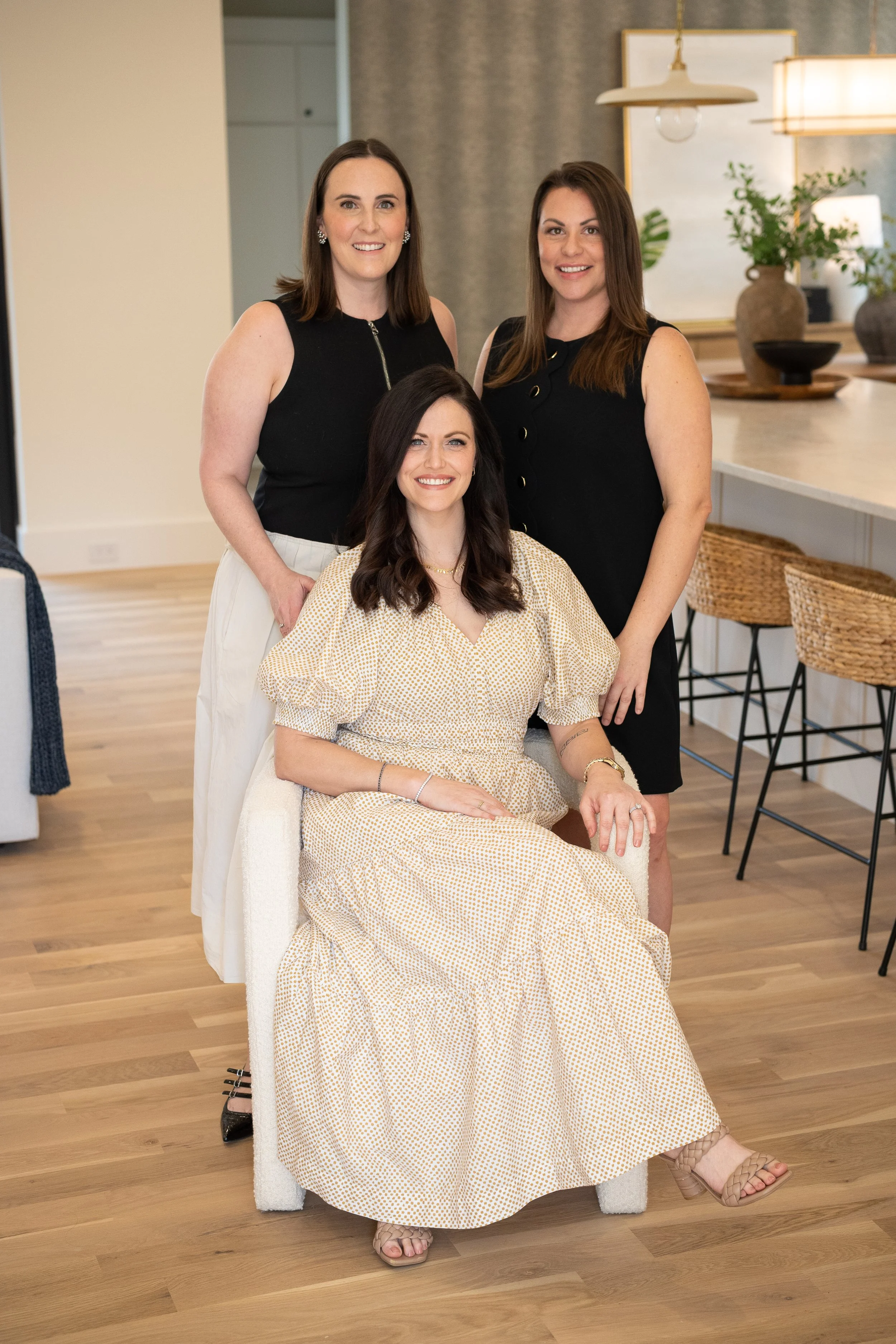 Three women posing for a photo in a modern, well-lit indoor space with wooden floors and bar stools. One woman is seated in a light-colored dress, and two women are standing behind her, all smiling.