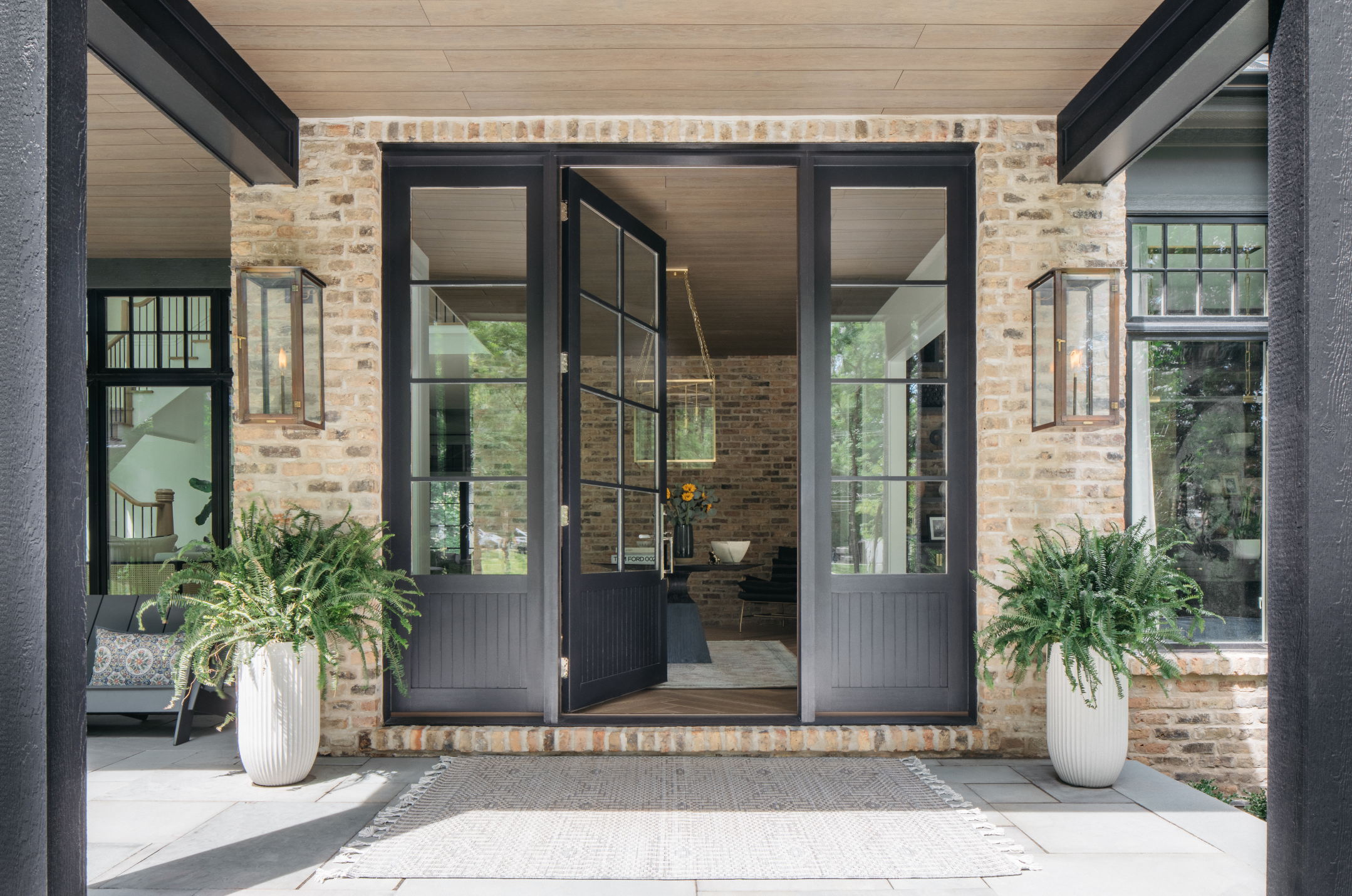 Open black-framed glass door leading into a brick-walled interior with a vase of sunflowers and modern furniture, flanked by two potted ferns outside on a tiled porch with a rug.