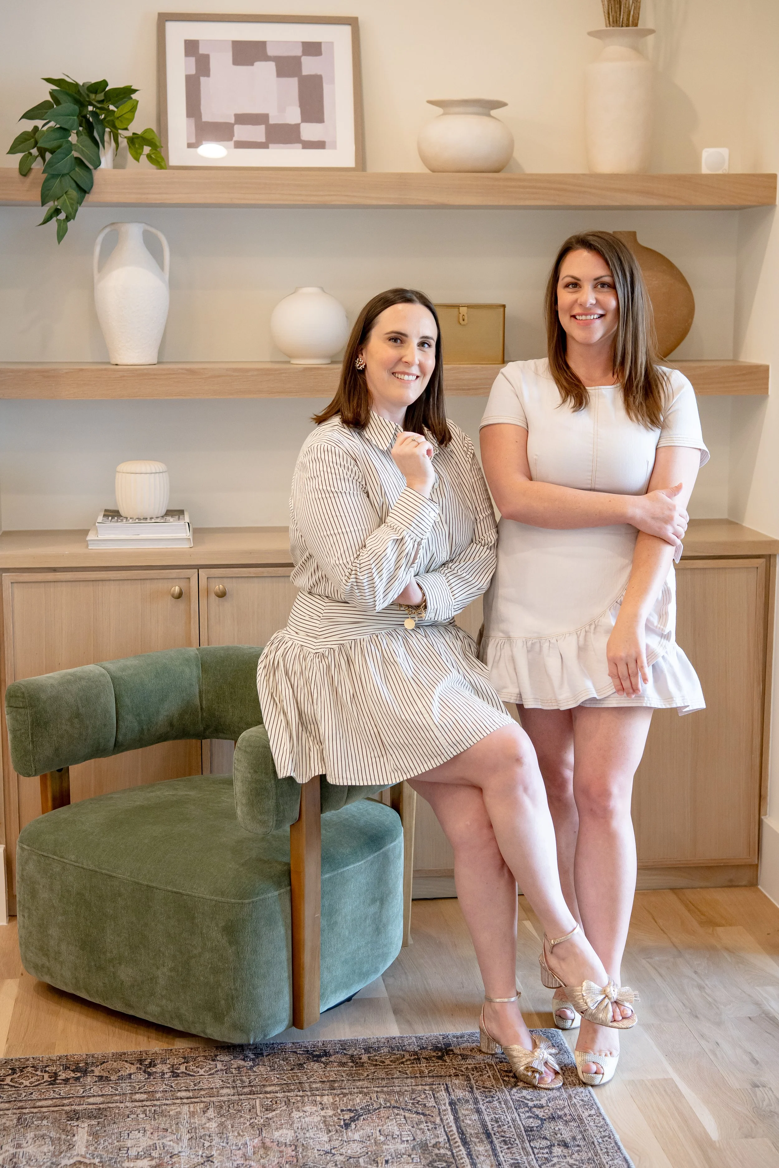 Two women pose together in a modern living room, one seated on a green velvet chair and the other standing next to her, both smiling at the camera.