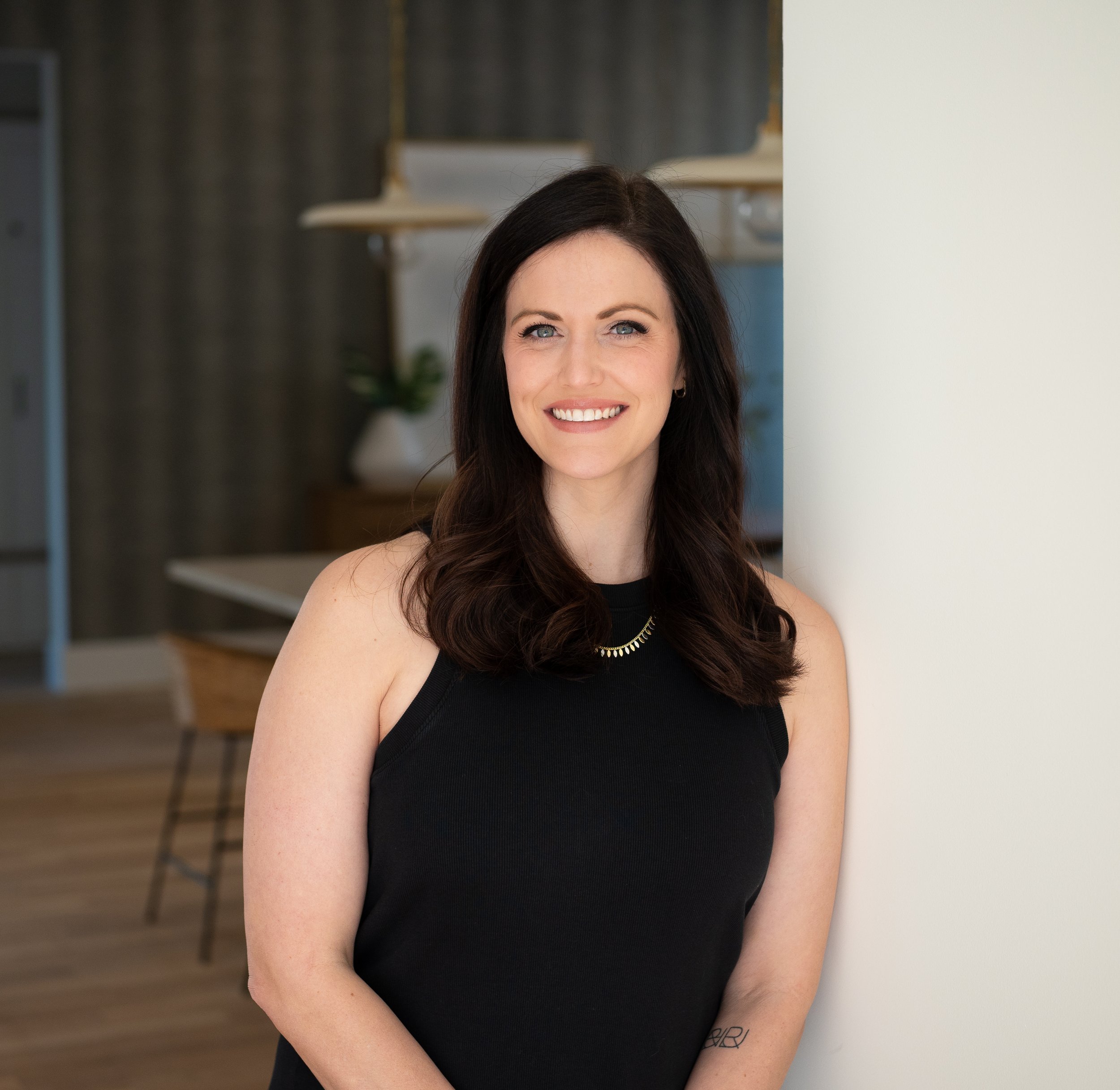 A woman with dark brown hair, wearing a sleeveless black top, smiling and leaning against a white wall in a modern indoor space.