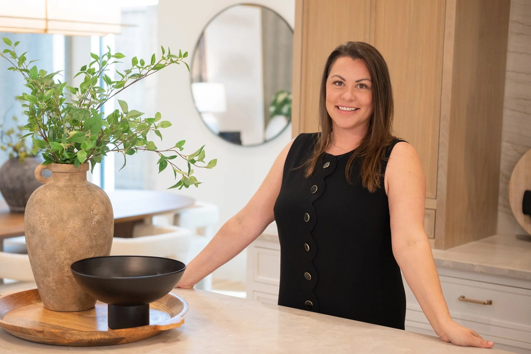 A woman with brown hair wearing a sleeveless black dress with gold buttons, smiling and standing in a modern kitchen, resting her hands on a white countertop, with a vase of greenery, a black bowl, and a wooden tray on the counter.