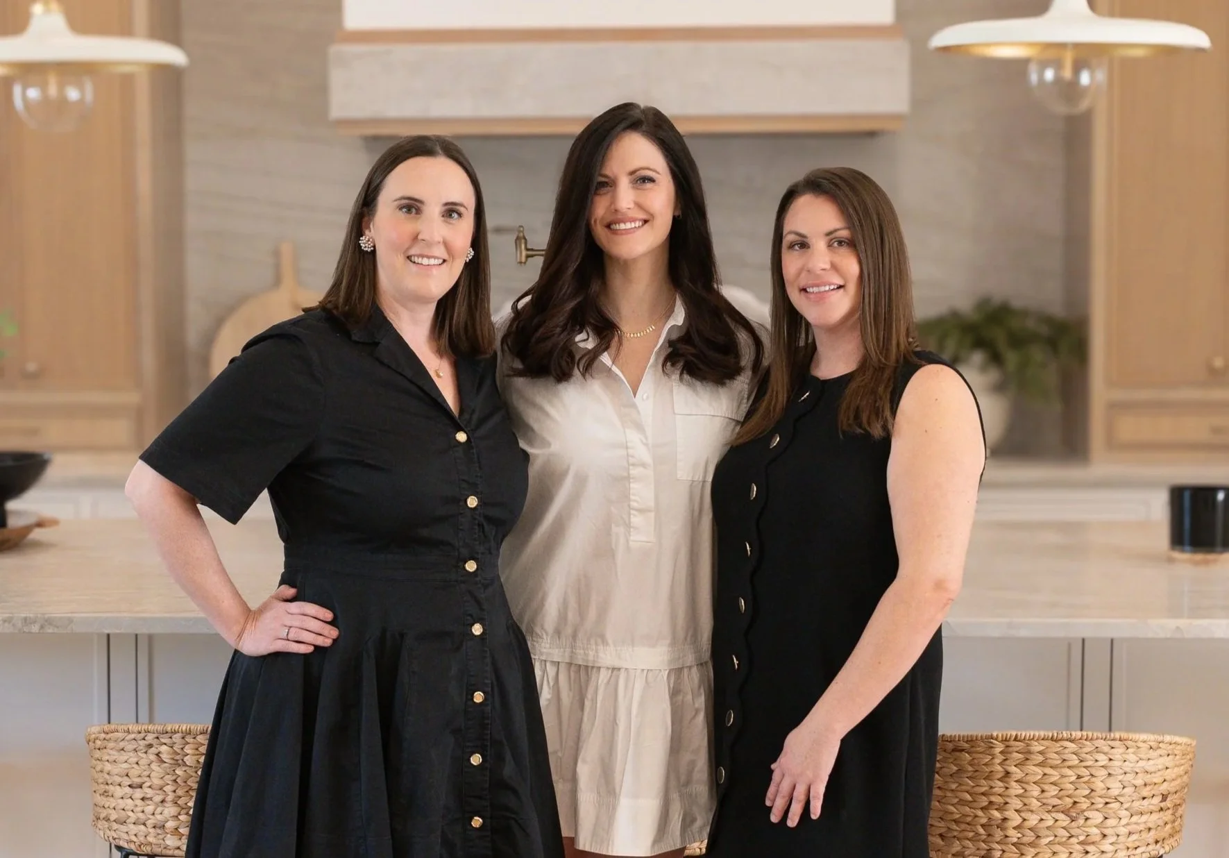 Three women standing in a kitchen, smiling at the camera, with wooden cabinets and a light-colored countertop in the background.