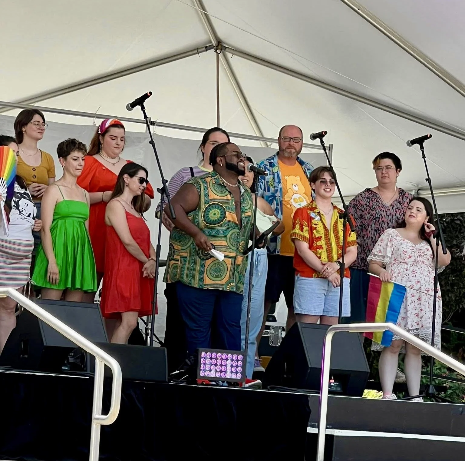 Group of diverse people performing on stage at an outdoor event, some wearing colorful casual clothing, one person holding a rainbow fan, and one holding a rainbow flag.