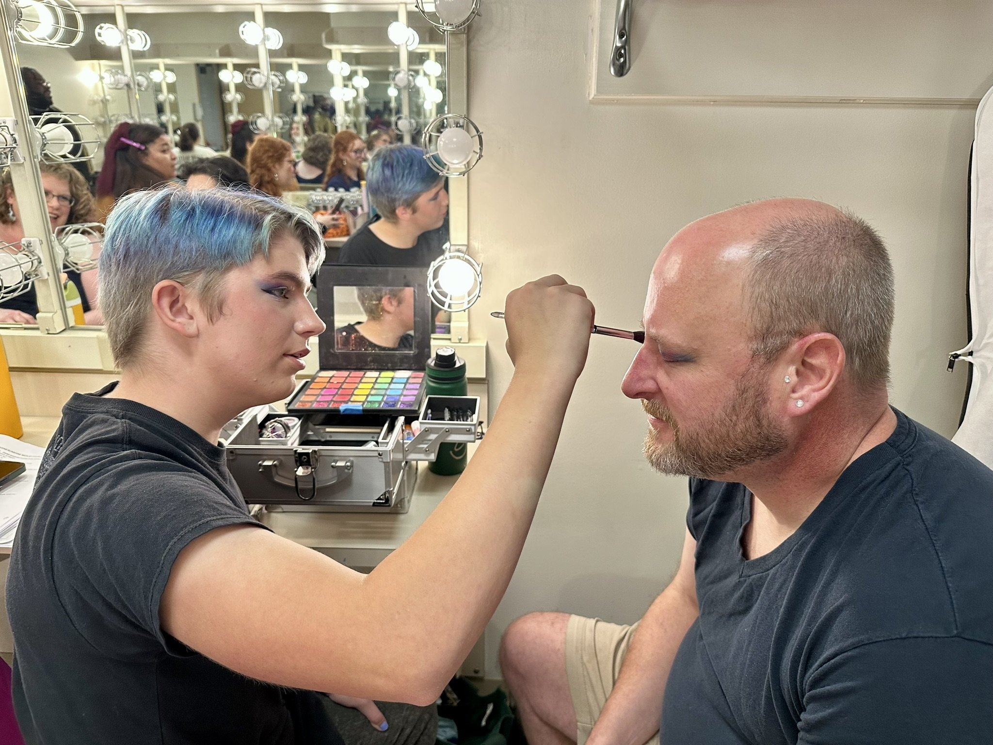 Makeup artist applying makeup to a man's face in a dressing room with mirrors and lights