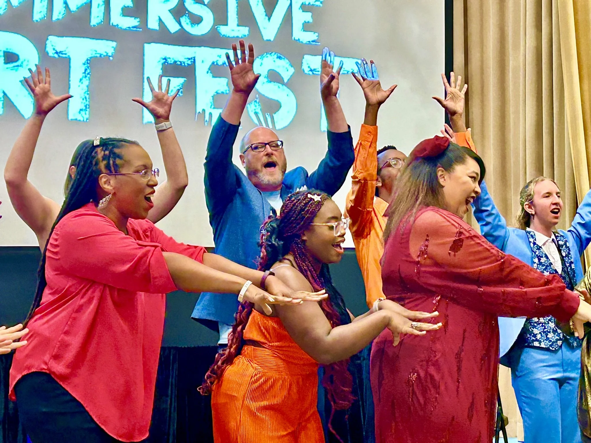 Group of people on stage with raised hands, singing, and smiling during a performance or celebration.