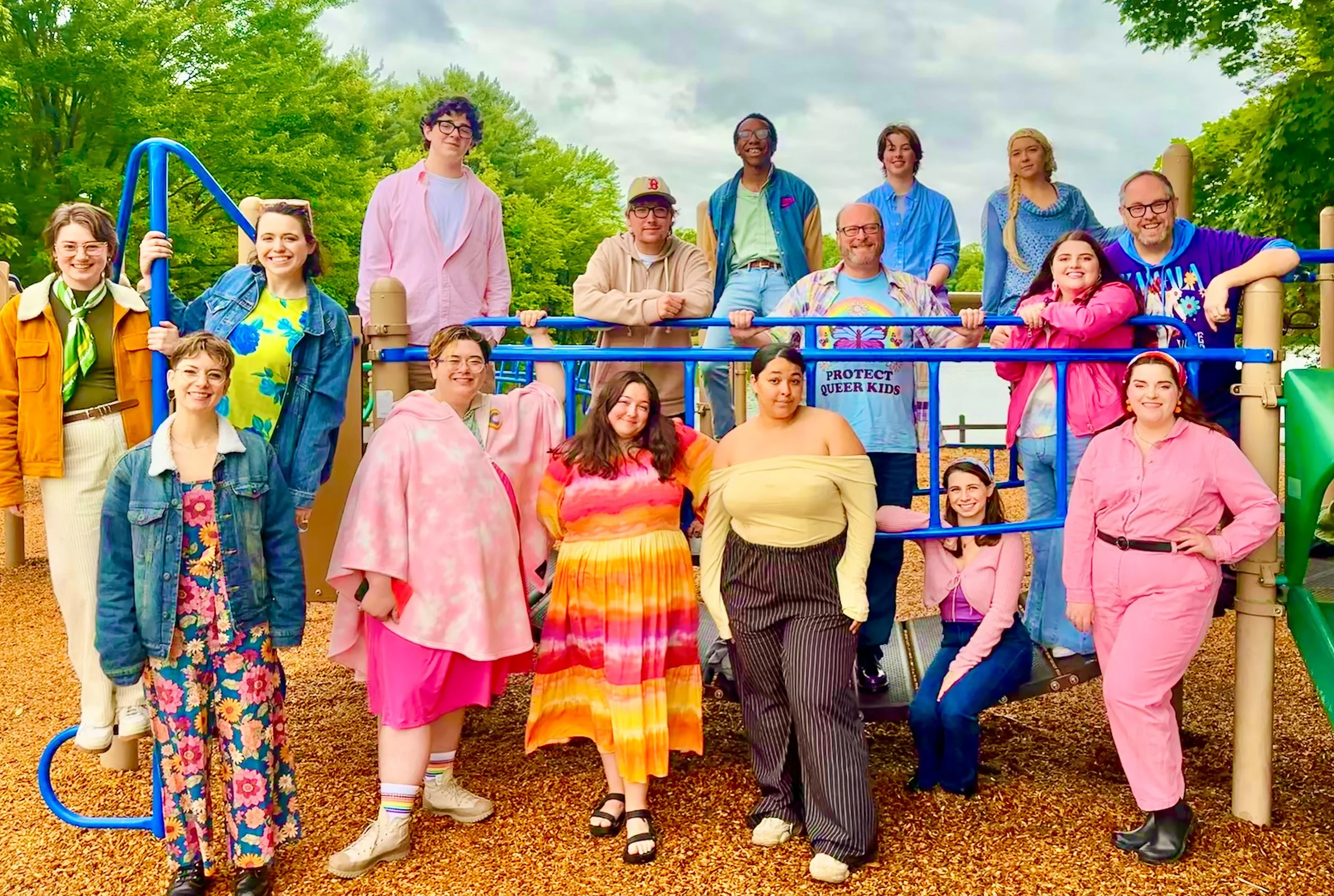 Group of diverse people, including children and adults, gathering on a playground structure outdoors, with trees and cloudy sky in the background.