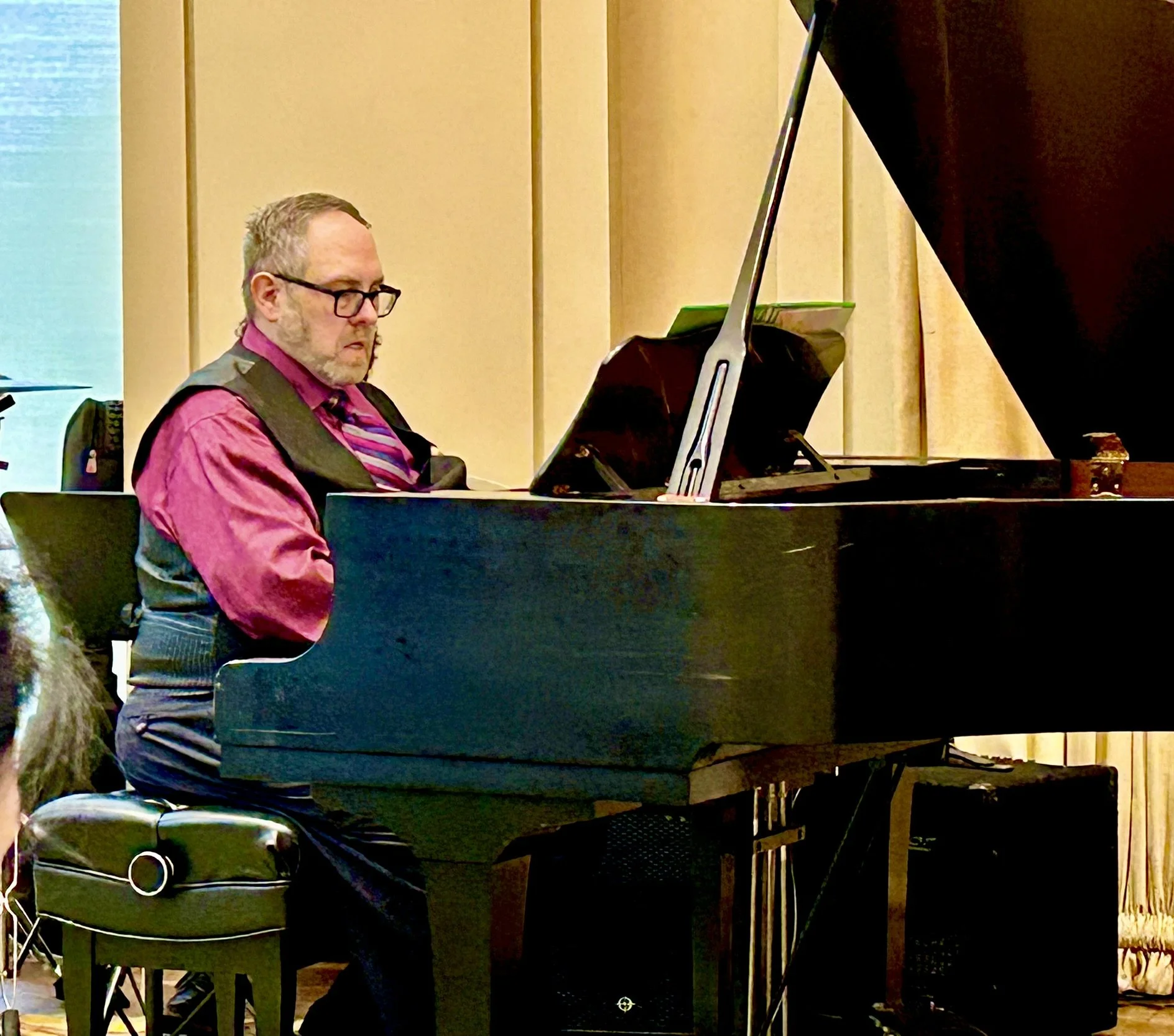 A man wearing glasses, a pink shirt, a vest, and a striped tie playing a grand piano during a performance in an indoor setting.