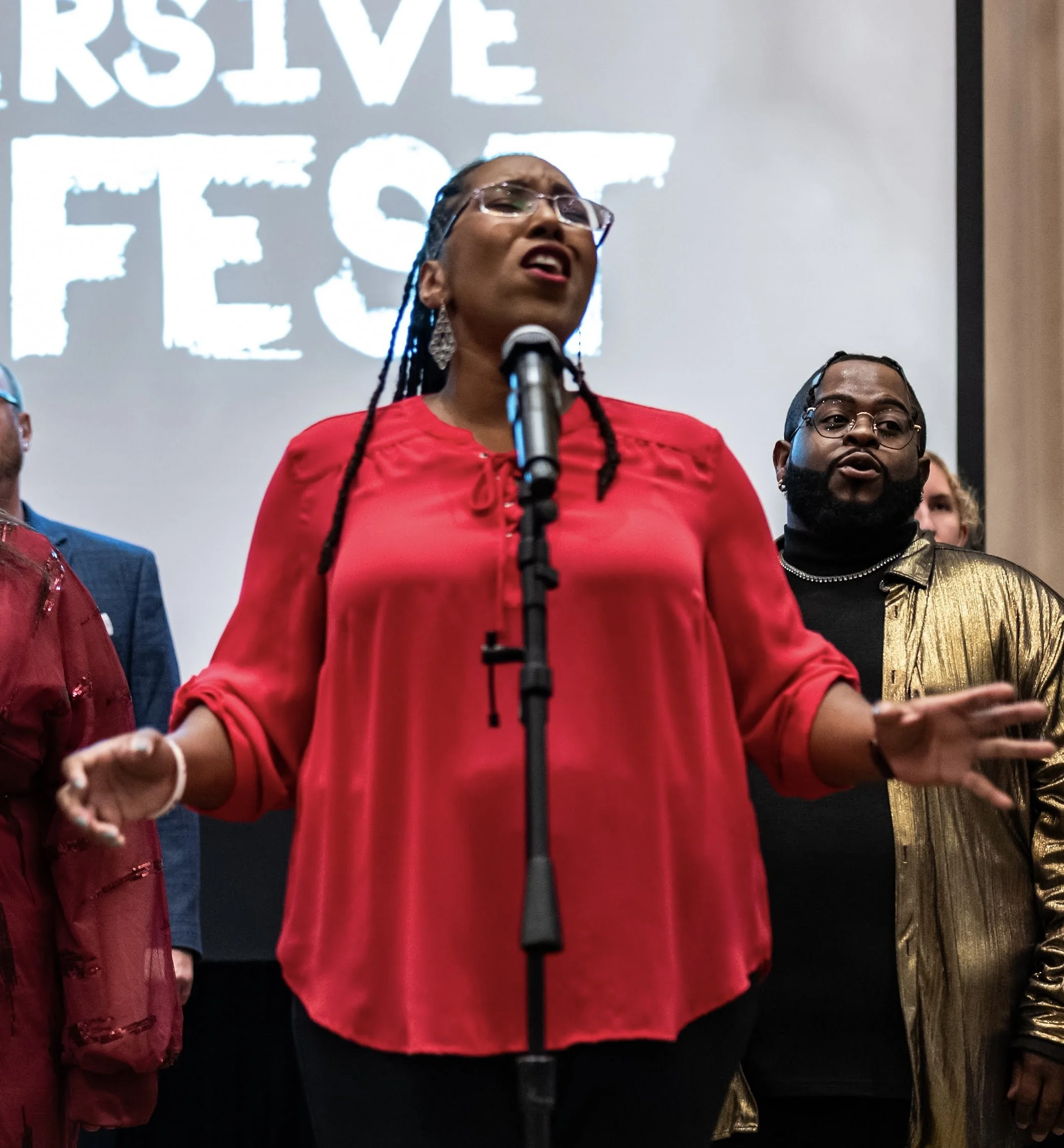 A woman in a red blouse is speaking or singing at a microphone during an event with a sign that reads 'RESERVE THE FESS' in the background, surrounded by other people.