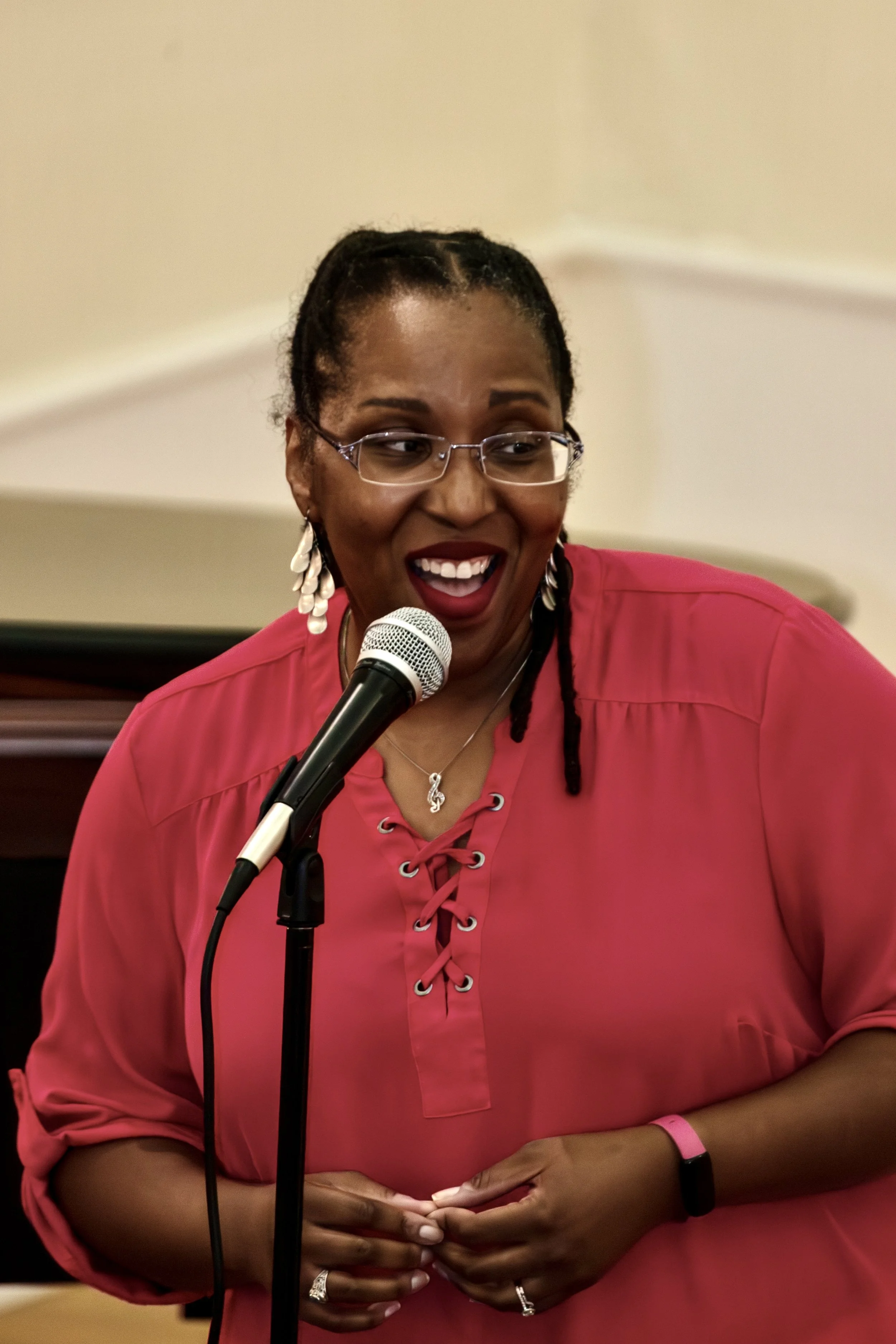 A woman speaking into a microphone, smiling, wearing glasses, a pink top, earrings, and a necklace, with a blurred background.