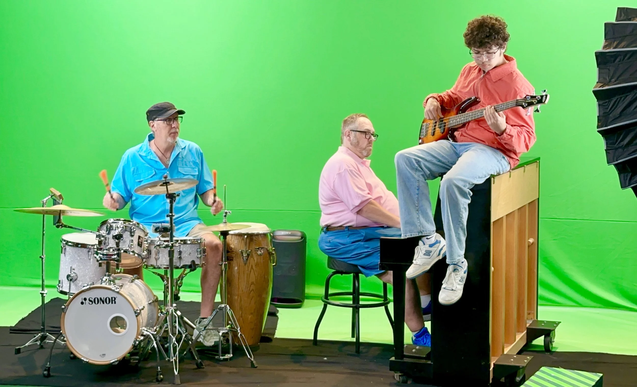 Three musicians performing in front of a green screen: a drummer, a pianist, and a guitarist sitting on a box, with studio equipment around.
