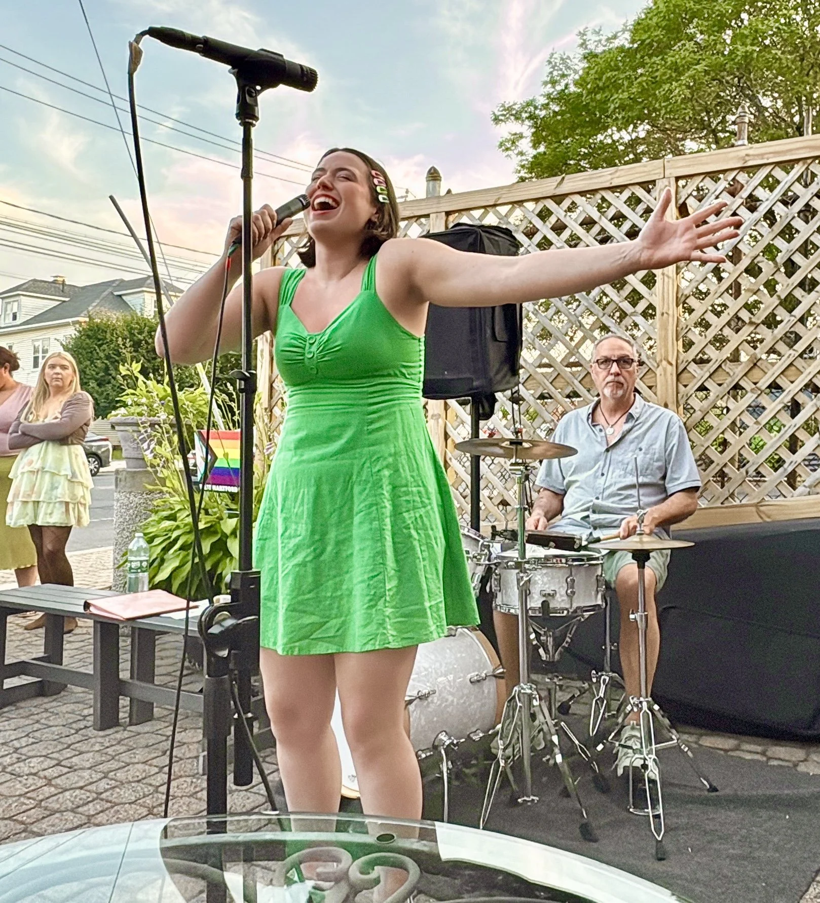 A woman singing into a microphone with outstretched arms at an outdoor event, accompanied by a drummer in the background, during daytime.