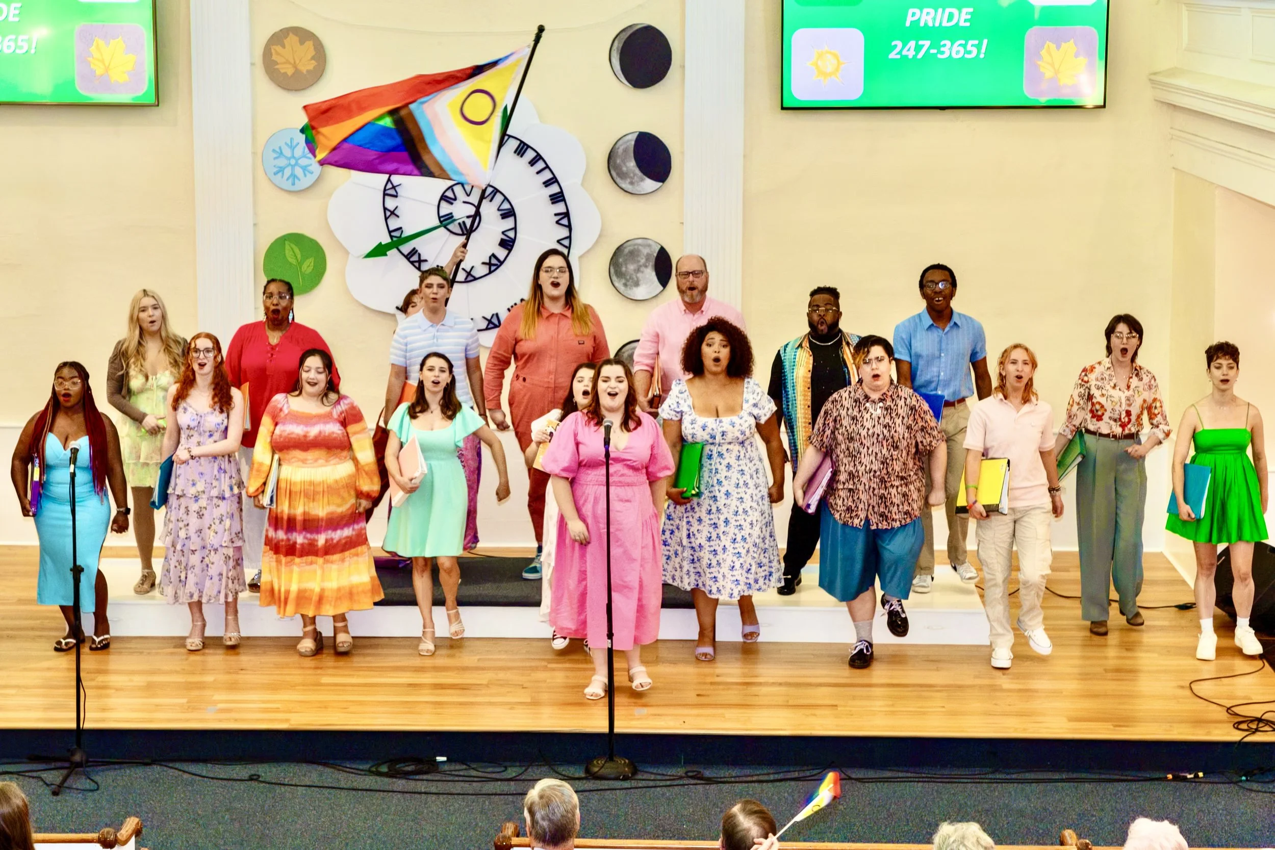 A diverse group of people singing or performing on a stage, with some holding folders or books, in a room decorated with a rainbow flag, a large clock, and moon phase symbols, and two digital screens displaying the word 'PRIDE' and the number '247-36