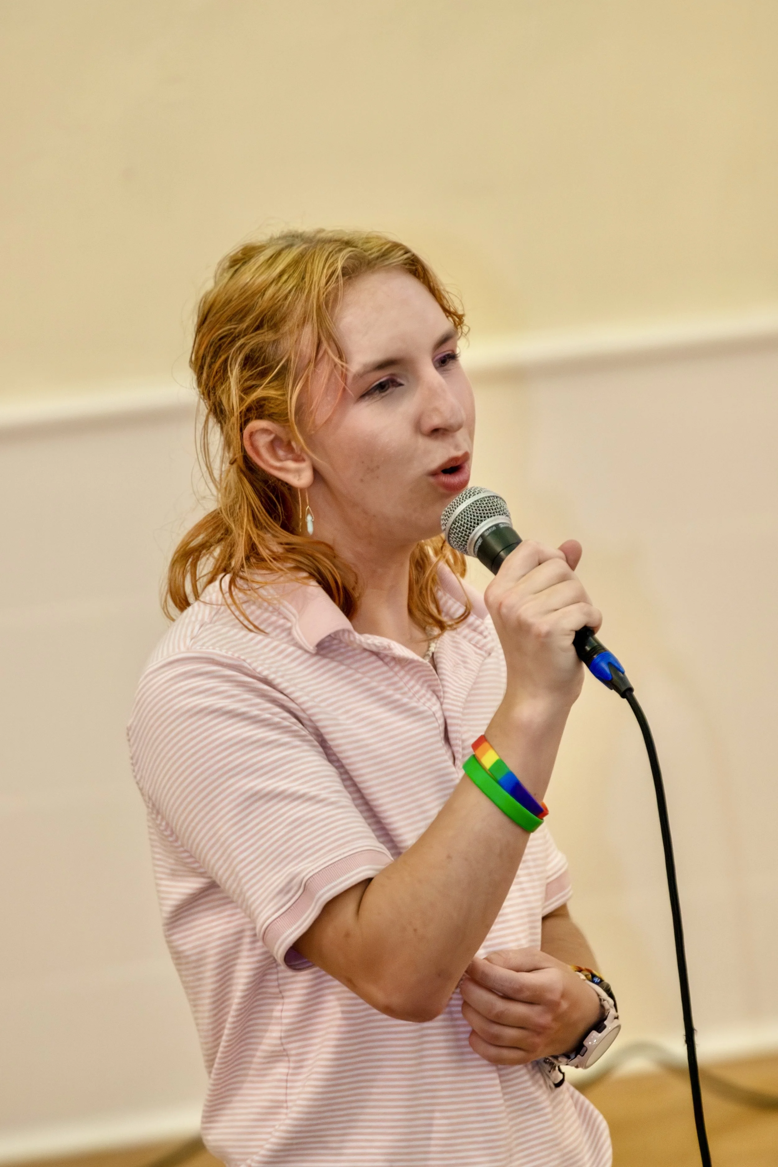 A young woman with red hair wearing a pink striped polo shirt and rainbow-colored wristbands, speaking into a microphone at an indoor event.